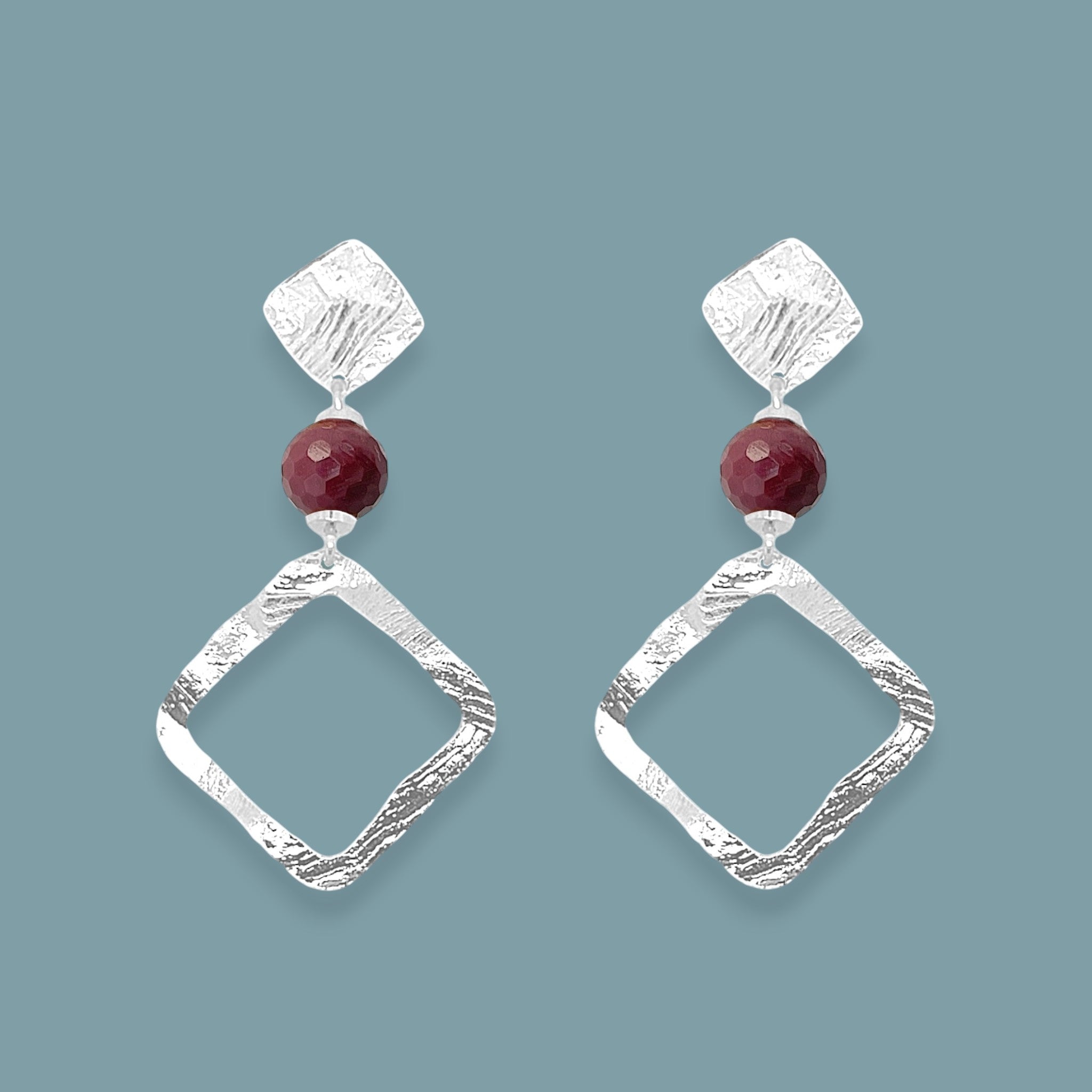 Boucles d'oreilles losanges sur puces en métal martelé argenté et perles de Mokaïte bordeaux, sur fond bleu gris