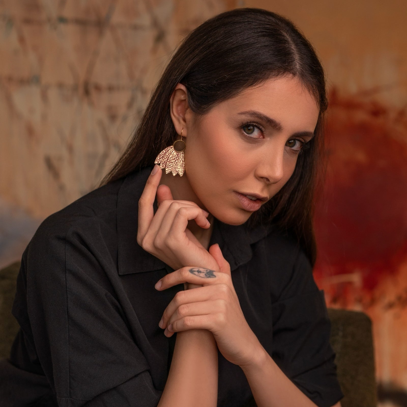 Grandes boucles d'oreilles dorées en forme de couronne de plumes en cuir or rosé froissé, portées à l'oreille par une femme brune