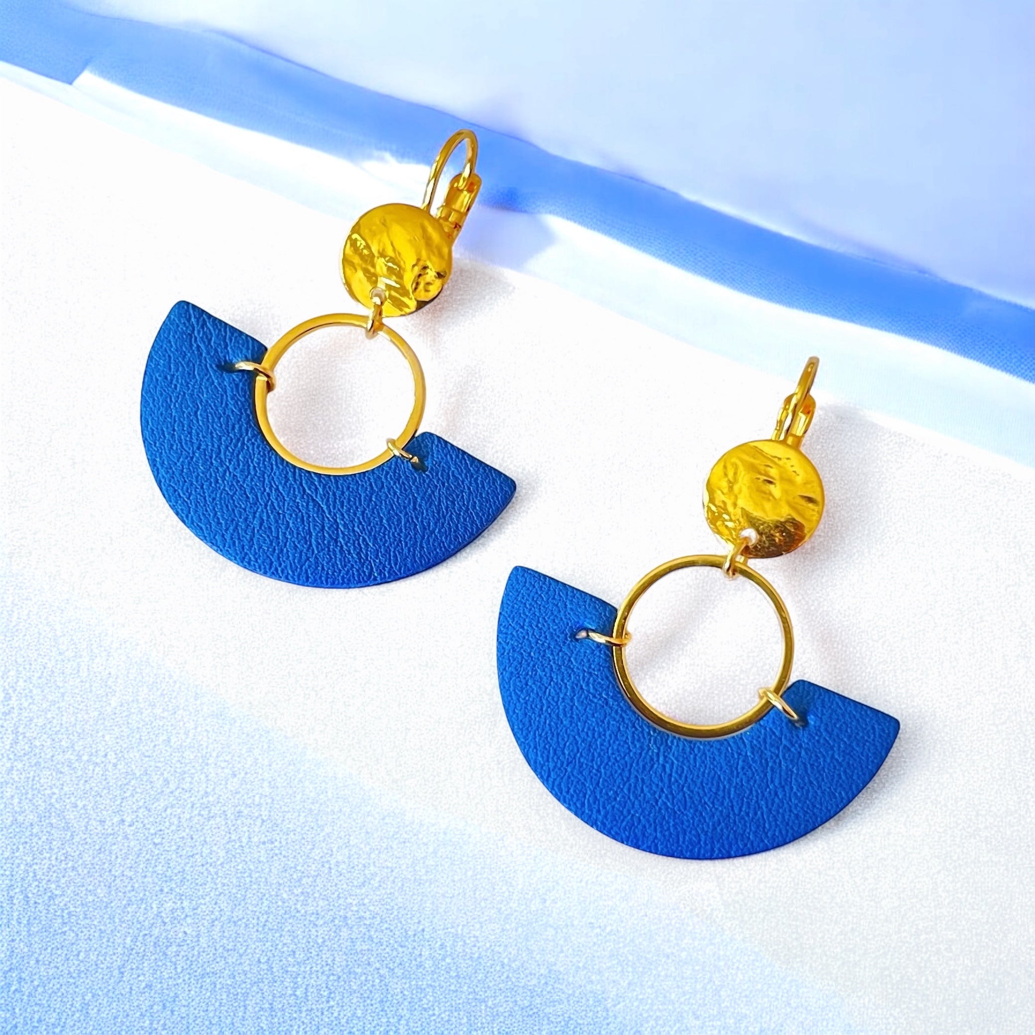 paire de boucles d'oreilles dorées pendantes en forme de demi-lunes en cuir bleu roi, sur fond blanc rayé bleu