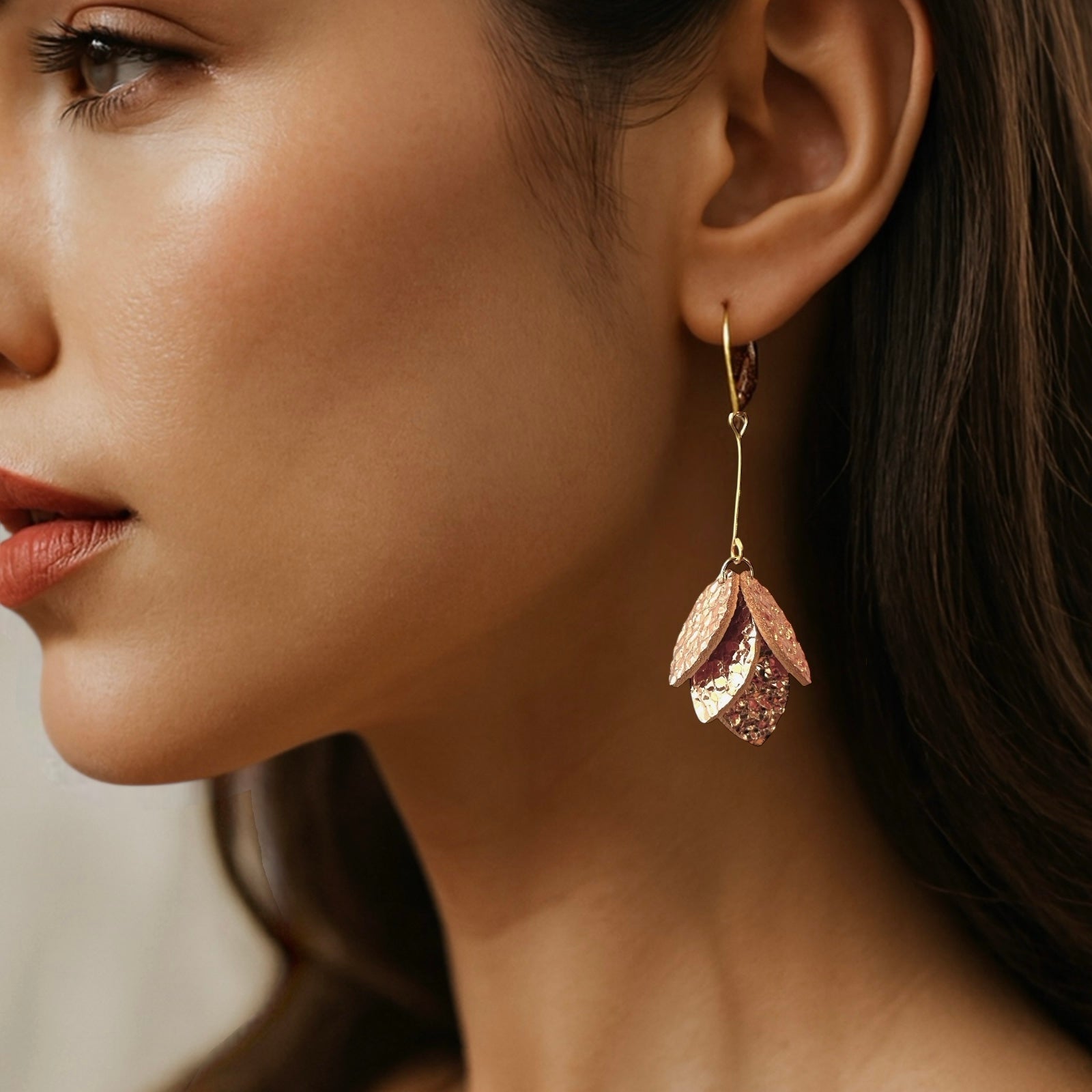 Boucles d'oreilles dorées longues et fines terminées par une grappe de pétales de cuir or rosé froissé, portées à l'oreille par une femme brune