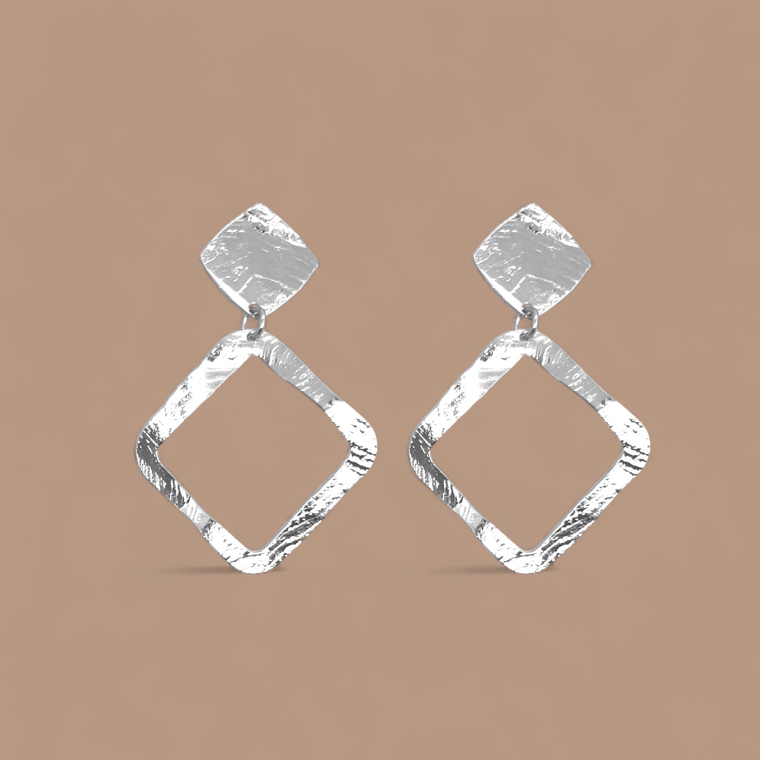 Boucles d'oreilles puces argentées en métal martelé avec pendentif en forme de losange, sur fond beige