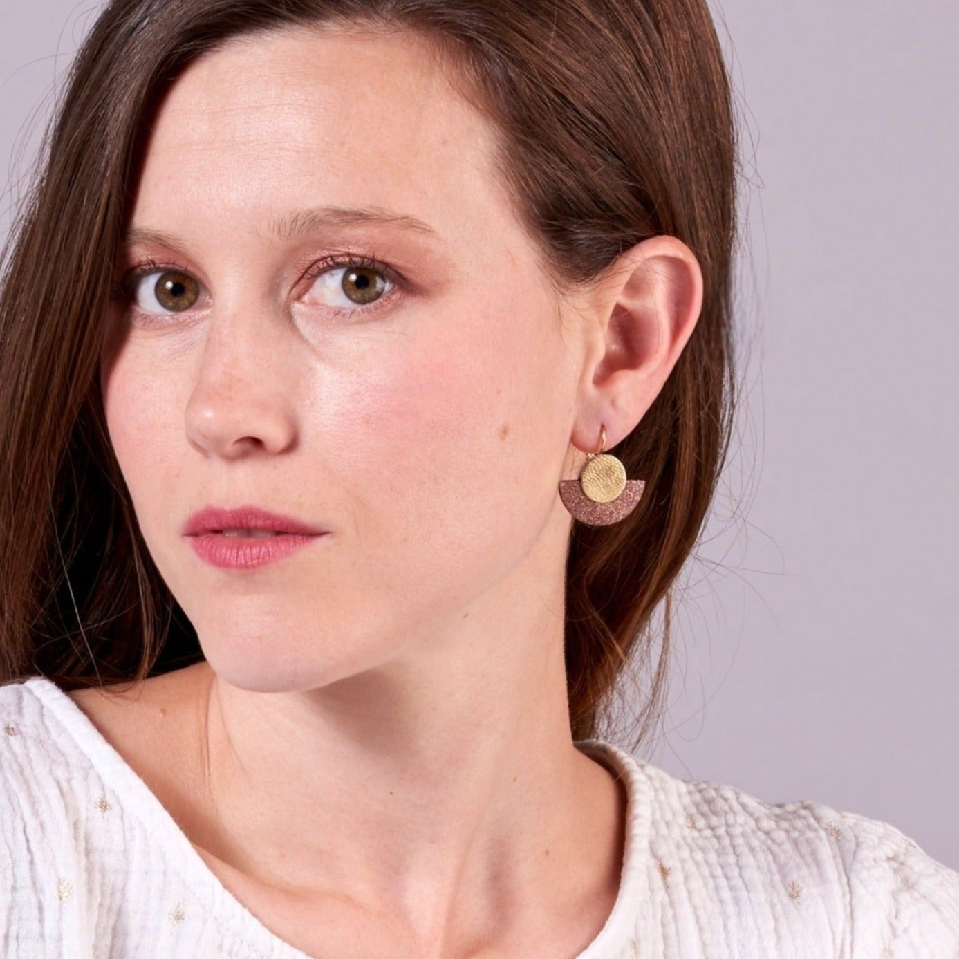 Jeune femme portant les boucles d'oreilles Téthys pailleté dorées en forme de demi-lunes en cuir vieux rose pailleté or et or recyclé, sur fond mauve