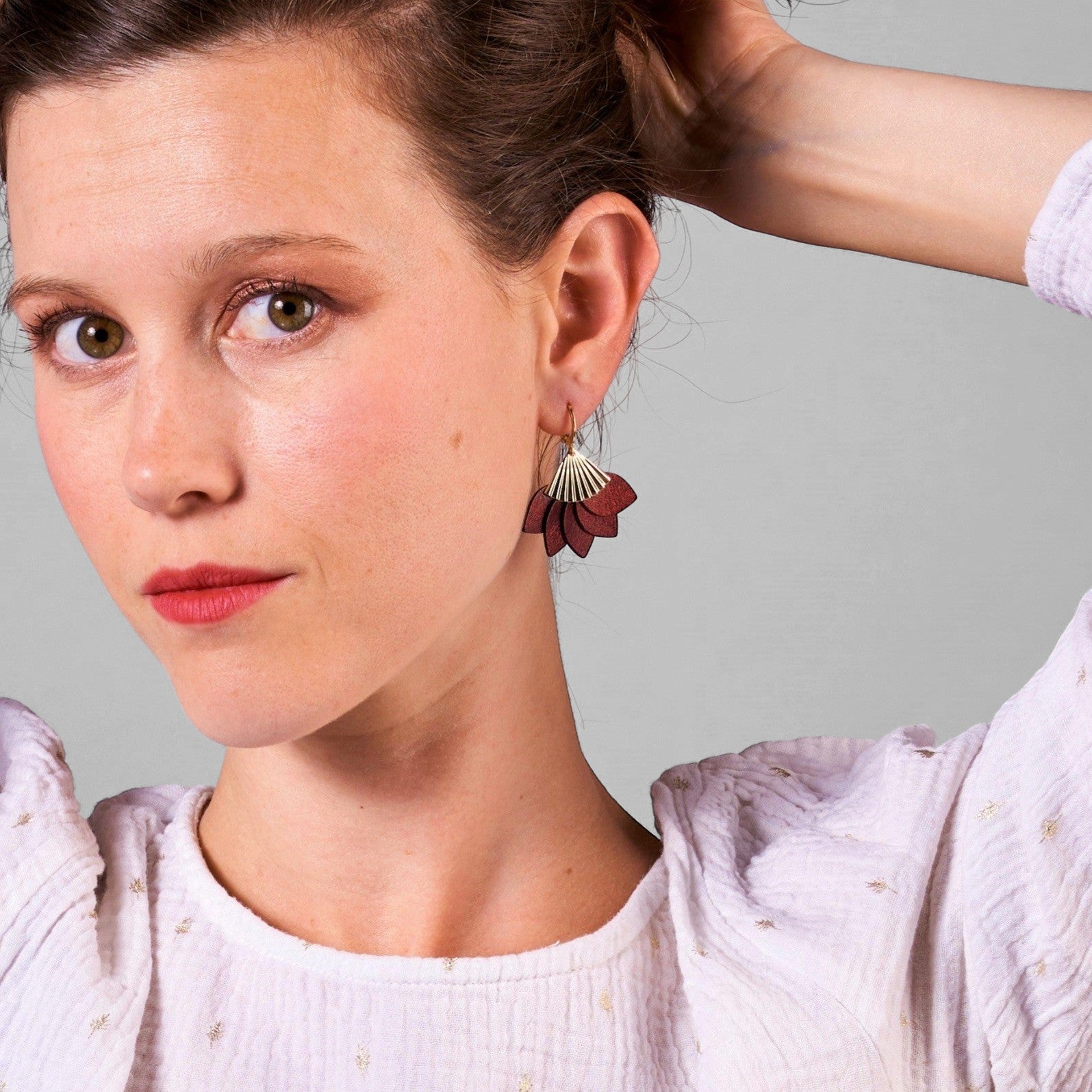 Jeune femme portant les boucles d'oreilles dorées en forme éventail en cuir recyclé grenat métallisé modèle Sakura, sur fond gris