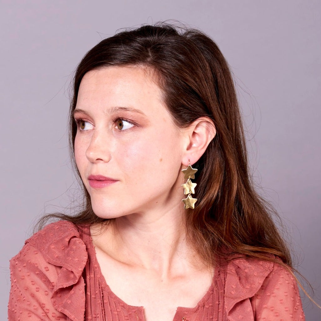 NUNE Boucles d'oreilles Starlight, chapelet d'étoiles en cuir recyclé doré, portées par une jeune femme aux cheveux longs bruns, vue de profil