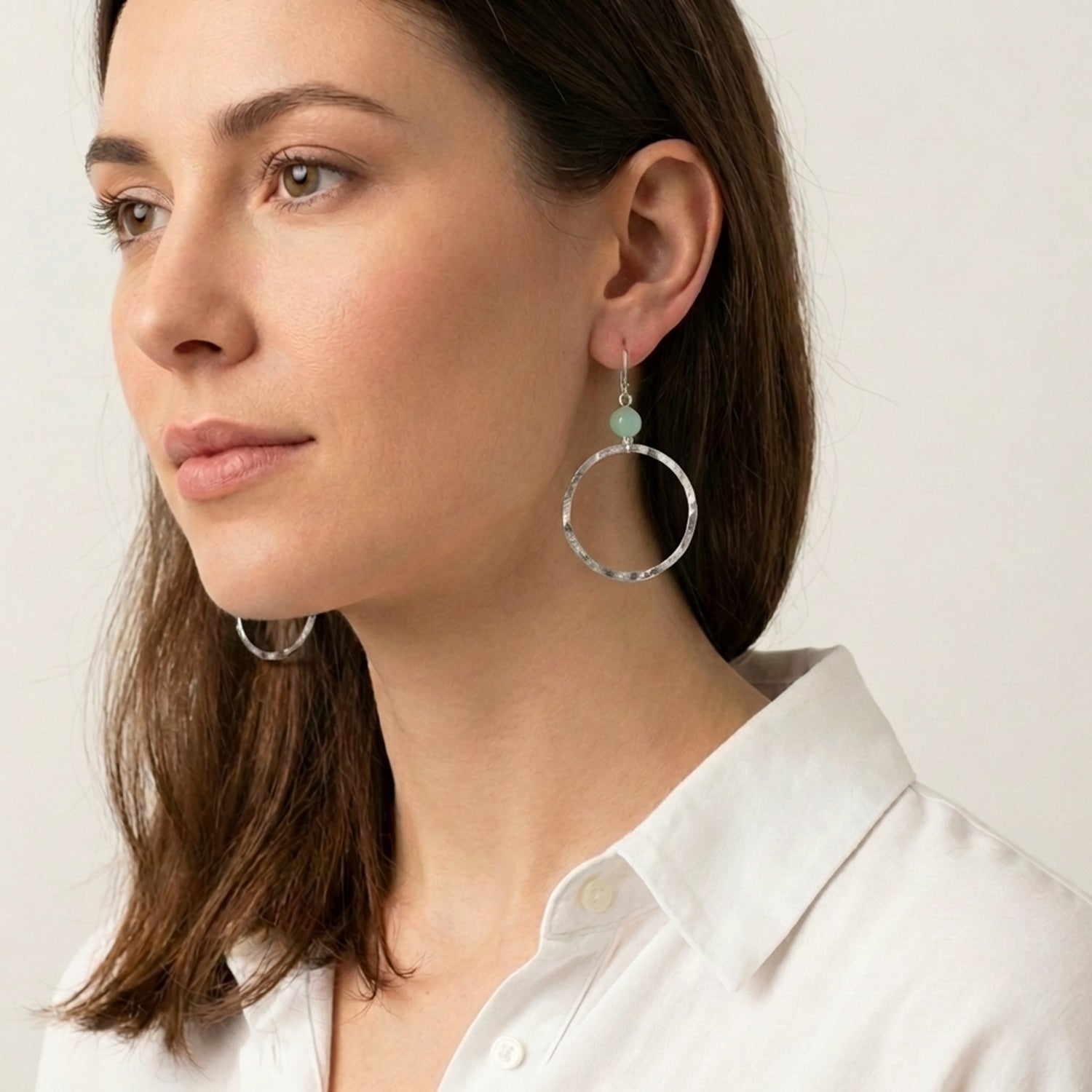 Boucles d'oreilles créoles de forme ronde en métal martelé argenté et perles de fluorite vert d'eau, portées à l'oreille par une femme brune