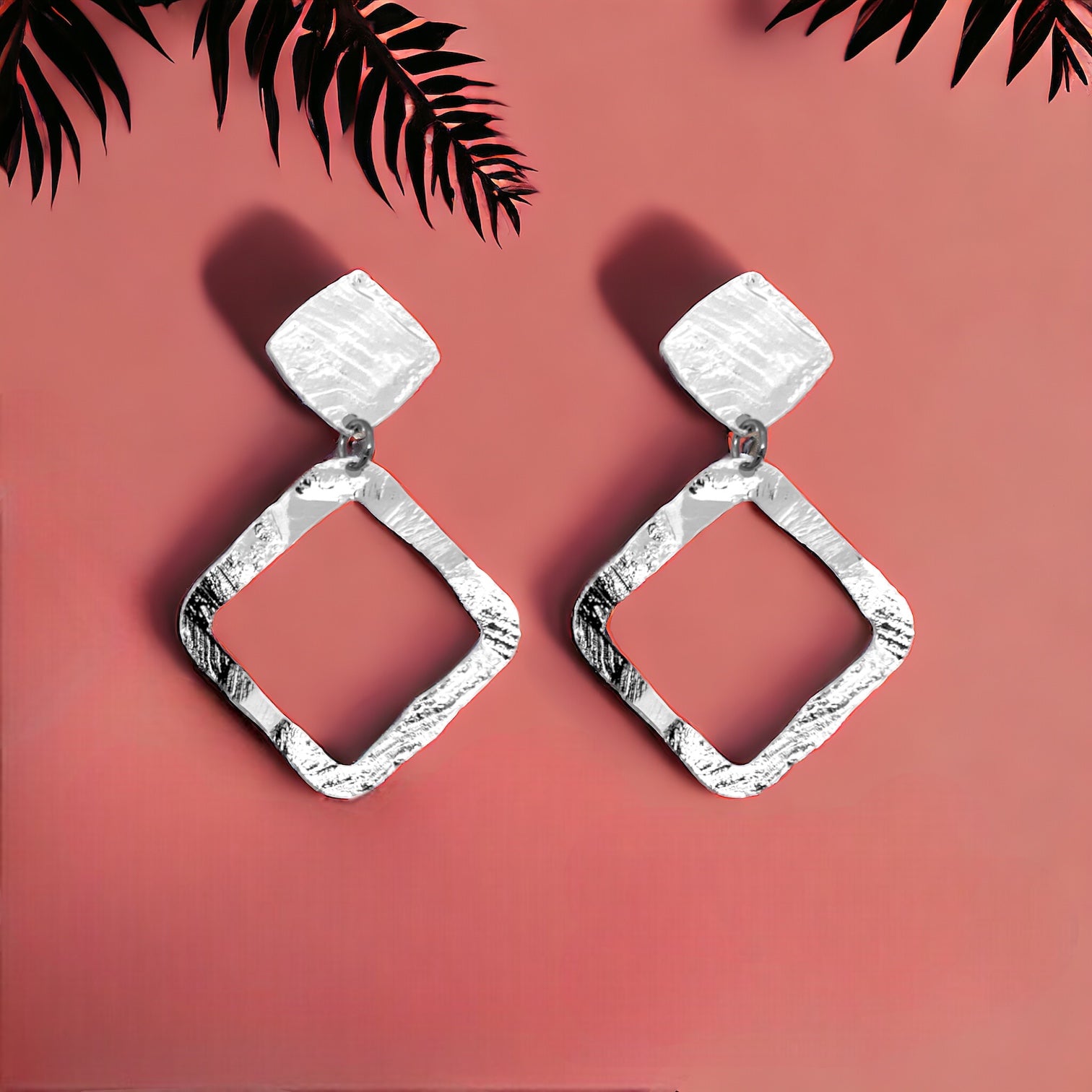Boucles d'oreilles puces argentées en métal martelé avec pendentif en forme de losange, sur fond rosé avec des feuilles de palmier