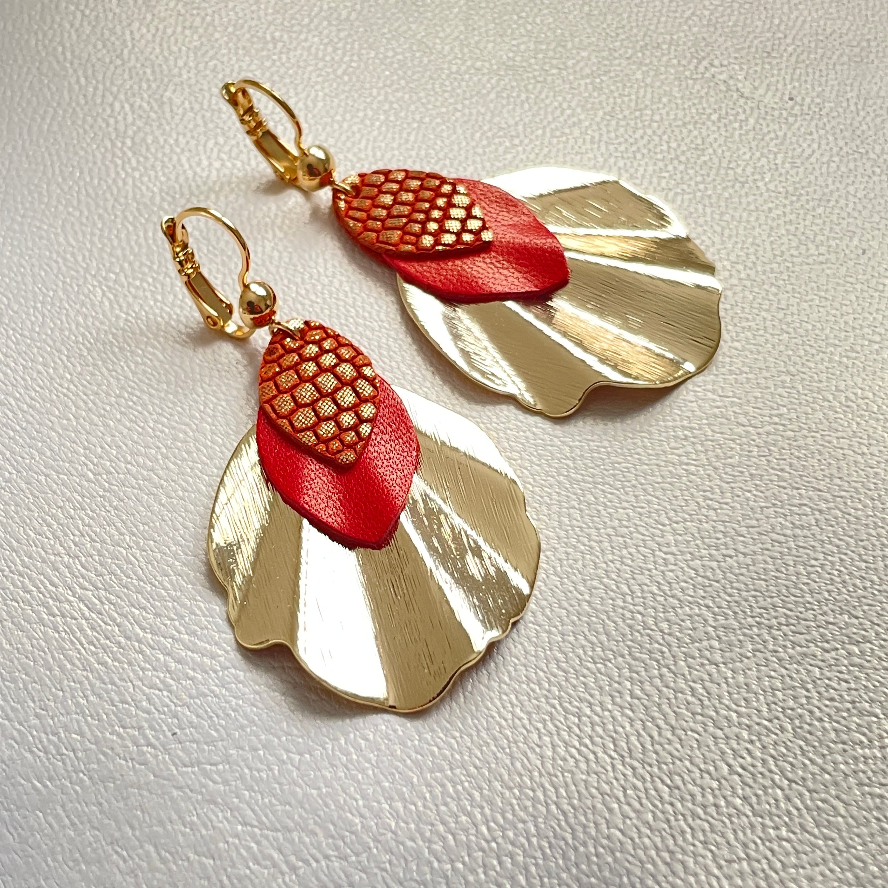 Boucles d'oreilles dorées Nune modèle Oko en forme de coquillages avec pétales de cuir rouge orangé nacré et orangé or façon serpent, sur fond cuir blanc cassé nacré