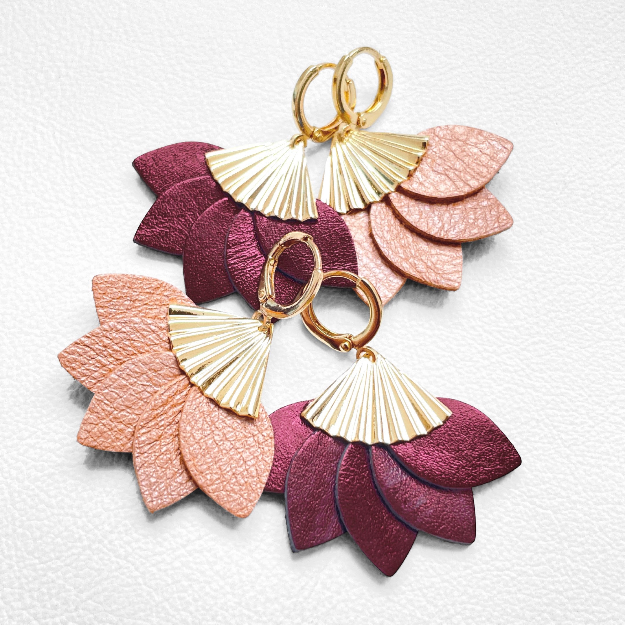 2 paires de boucles d'oreilles dorées en forme éventail en cuir recyclé corail nacré clair et grenat métallisé de la marque Nune modèle Sakura, sur fond de cuir blanc