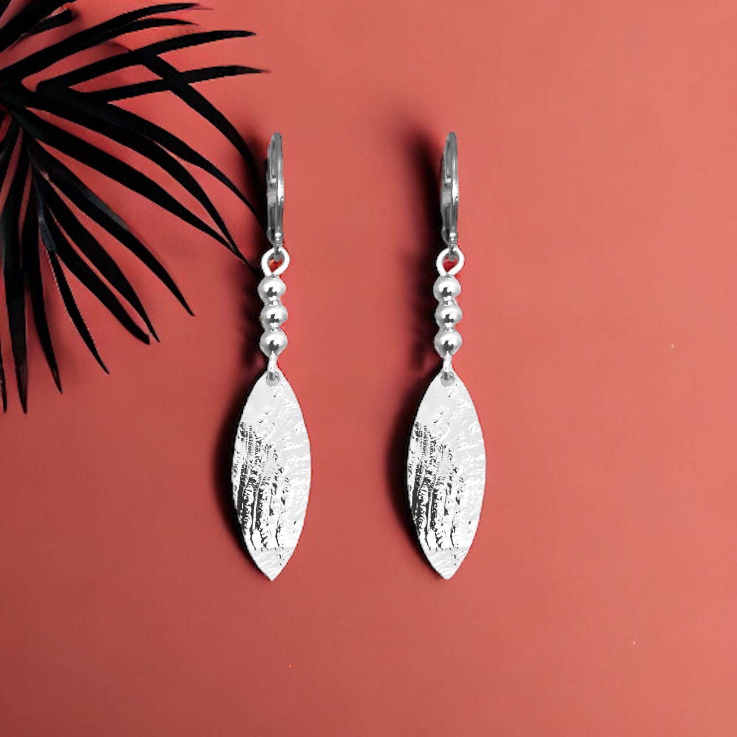 Boucles d'oreilles argentées fines pendantes longues en pointes, sur fond couleur brique avec feuille de palmier