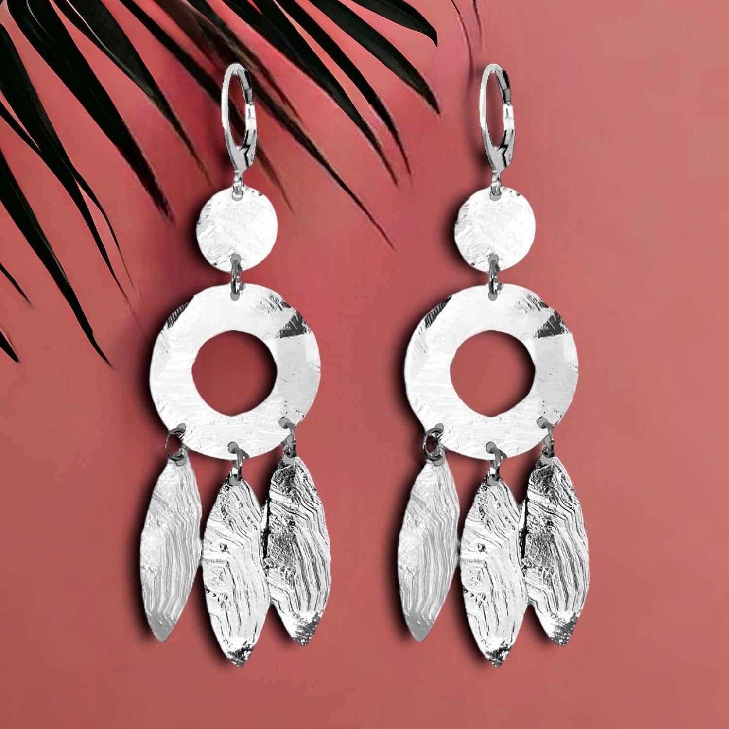Longues boucles d'oreilles argentées en métal martelé, de style ethnique bohème chic, avec leurs pampilles en forme de plumes, sur fond rosé avec une feuille de palmier