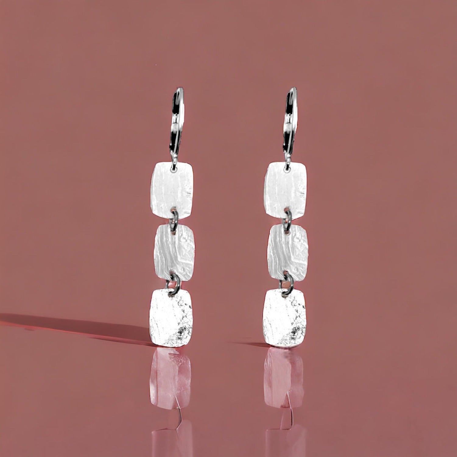 Fines boucles d'oreilles argentées pendantes en chapelet de 3 petits rectangles, sur fond marron clair