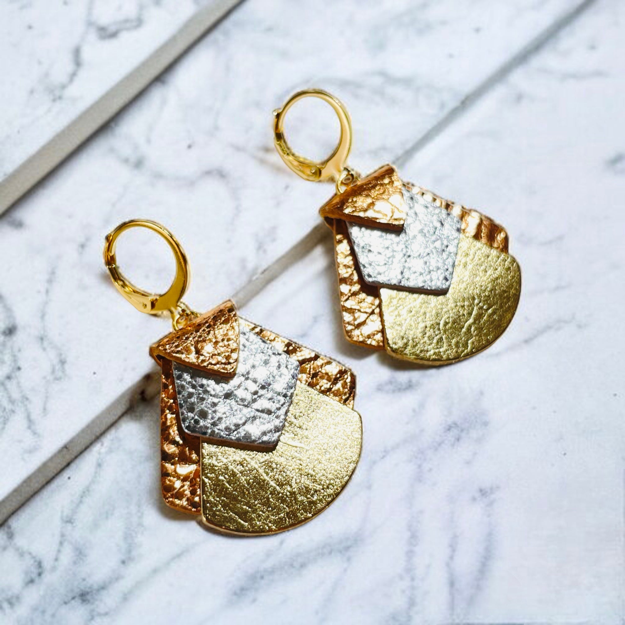 boucles d'oreilles aux formes géométriques en cuirs 3 ors, sur fond marbre blanc, vues de biais