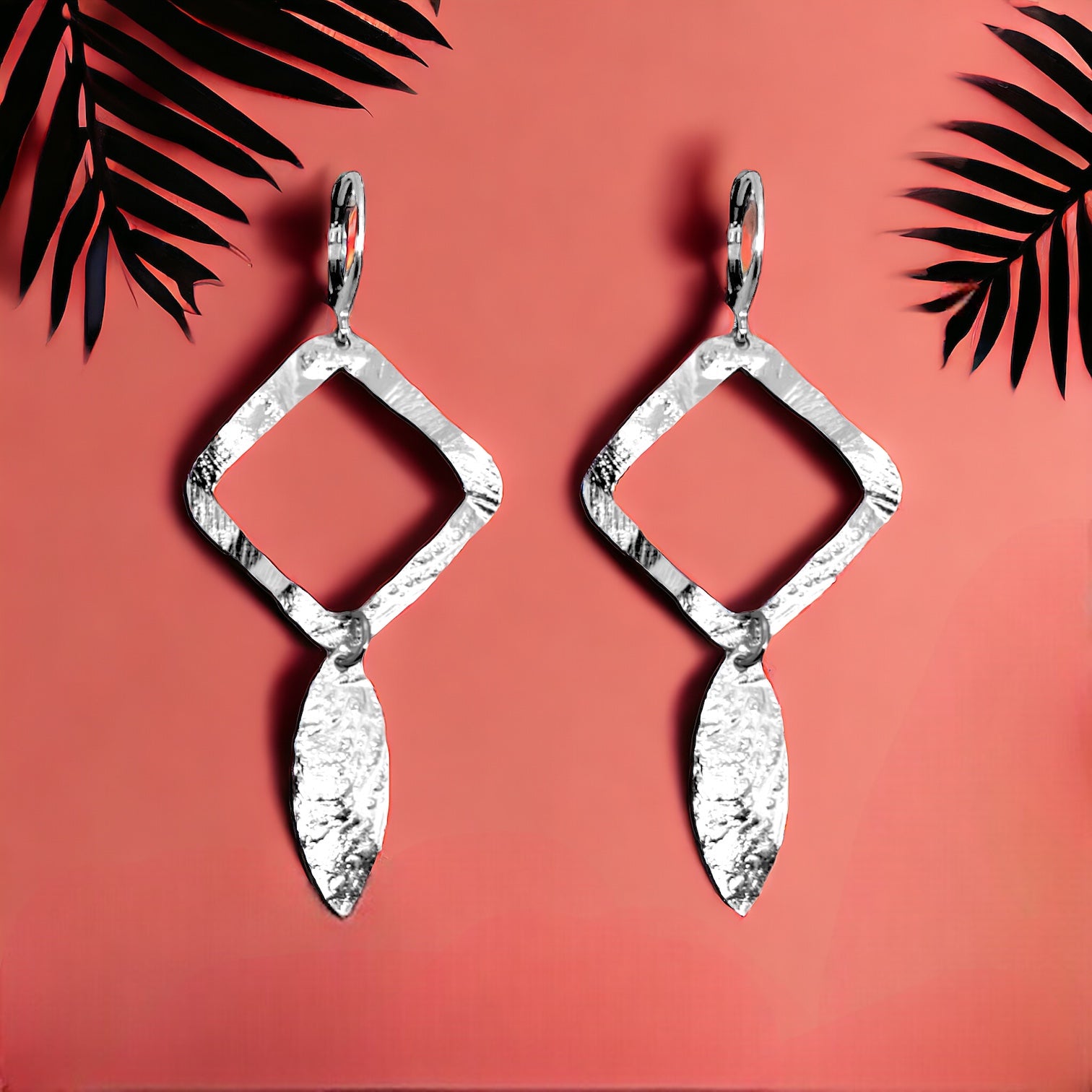Boucles d'oreilles argentées avec pendentif en forme de losanges et pointes, sur fond couleur brique avec feuilles de palmier