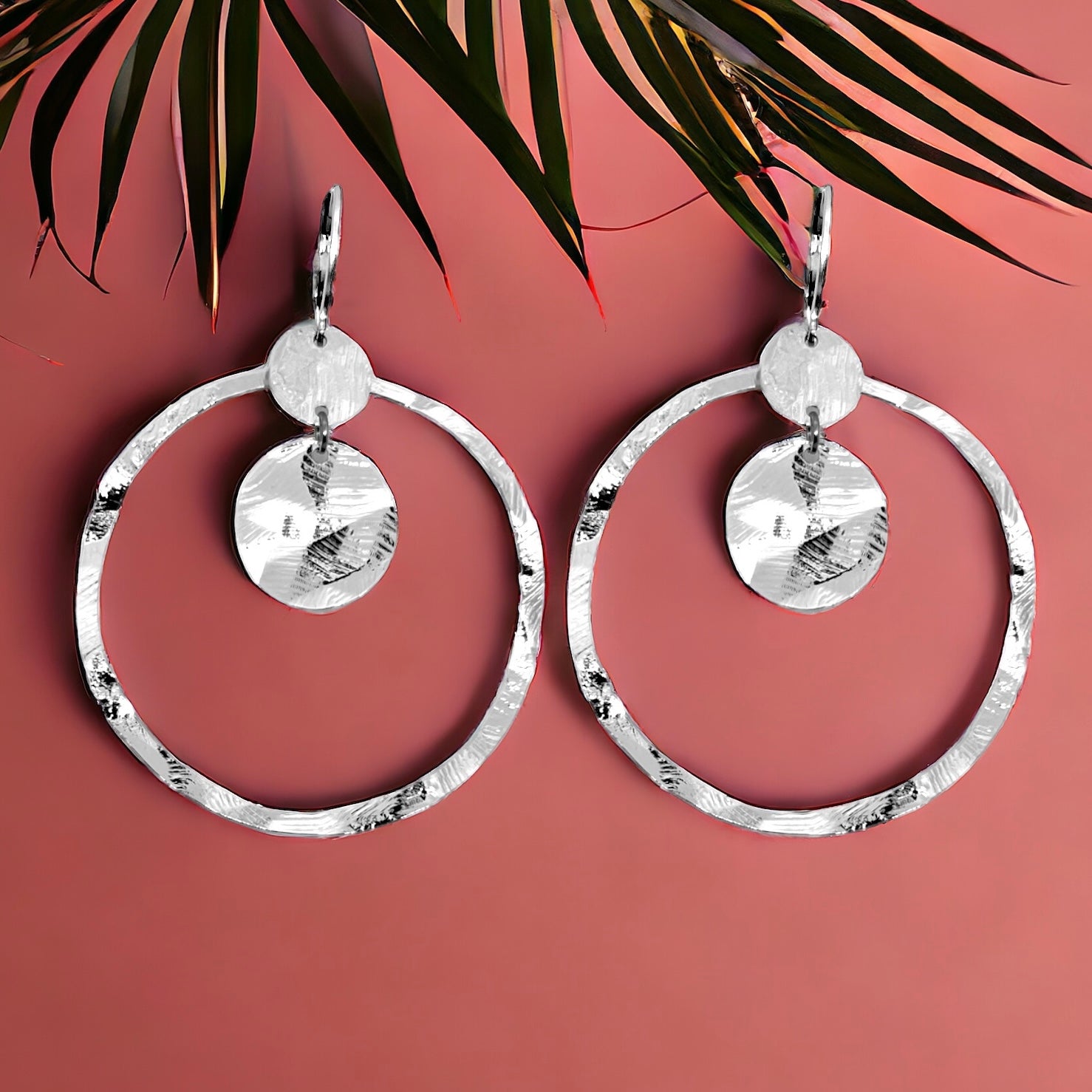 Grandes boucles d'oreilles créoles argentées avec sequins en métal martelé, avec attaches-dormeuses, sur fond rosé avec feuille de palme