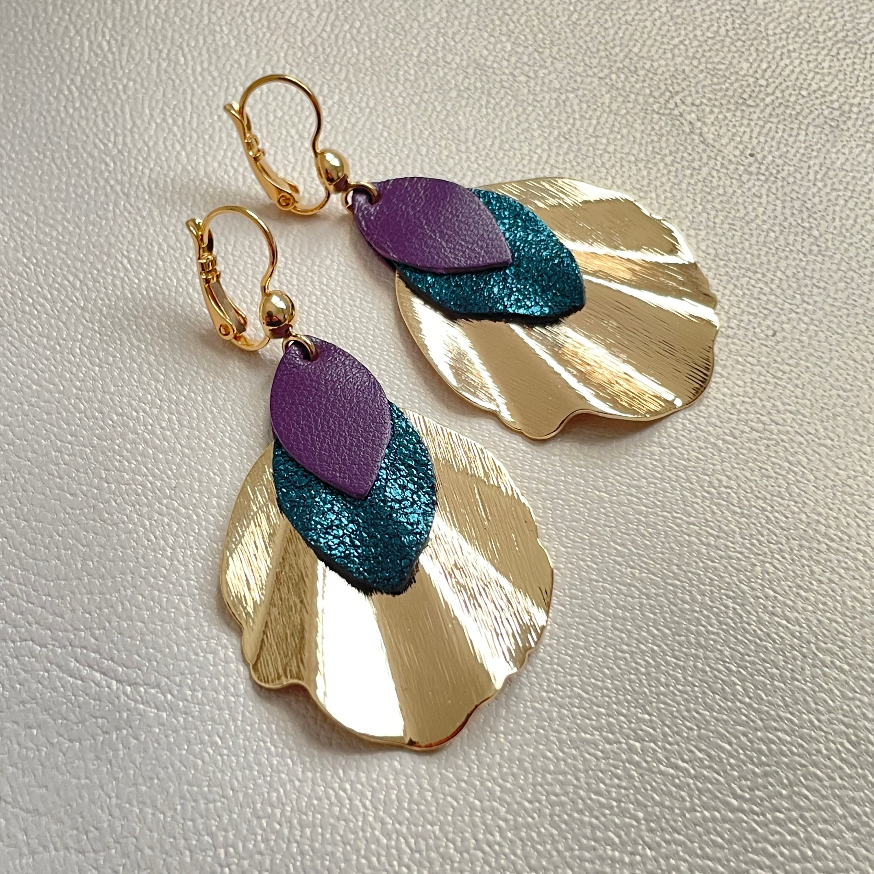 Boucles d'oreilles dorées Nune modèle Oko en forme de coquillages avec pétales de cuir bleu paon pailleté et violine satinée, sur fond cuir blanc cassé nacré