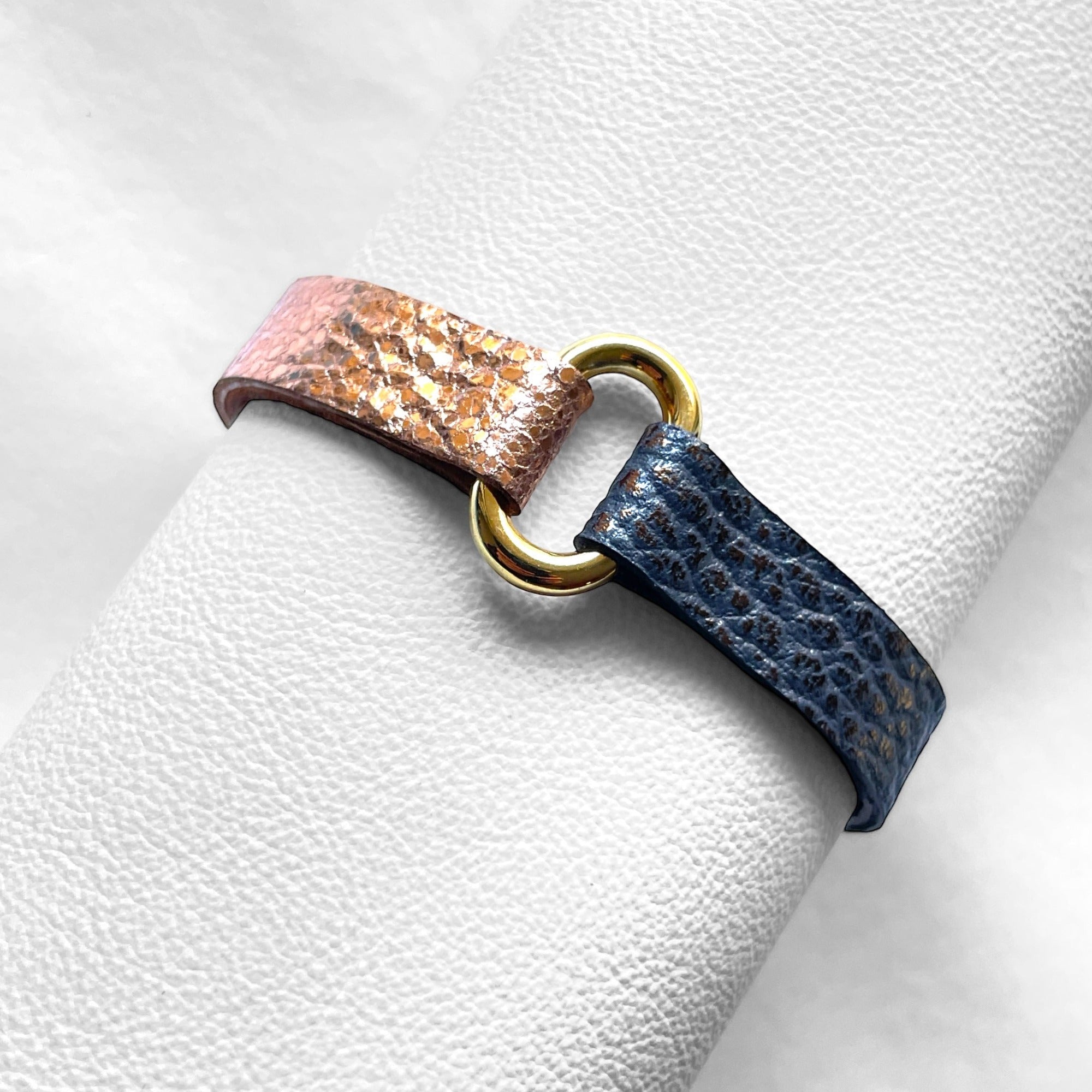 Bracelet bicolore à boucle dorée en cuirs recyclés or rose froissé et gris mordoré grainé, enroulé autour d'un rouleau de cuir blanc, vu de dessus