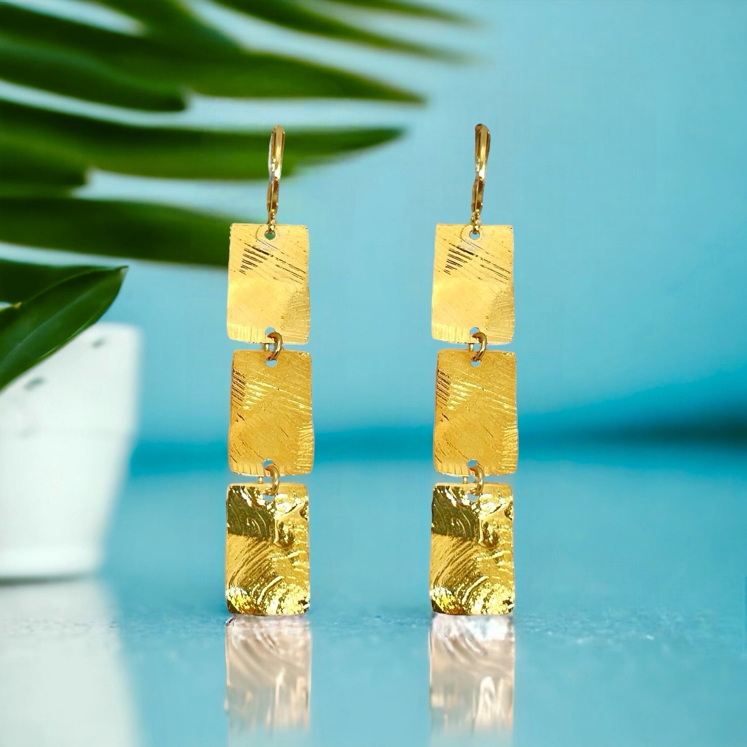 Boucles d'oreilles pendantes en chapelet de 3 rectangles en métal martelé doré à l'or fin, sur fond bleu et feuilles de palmier