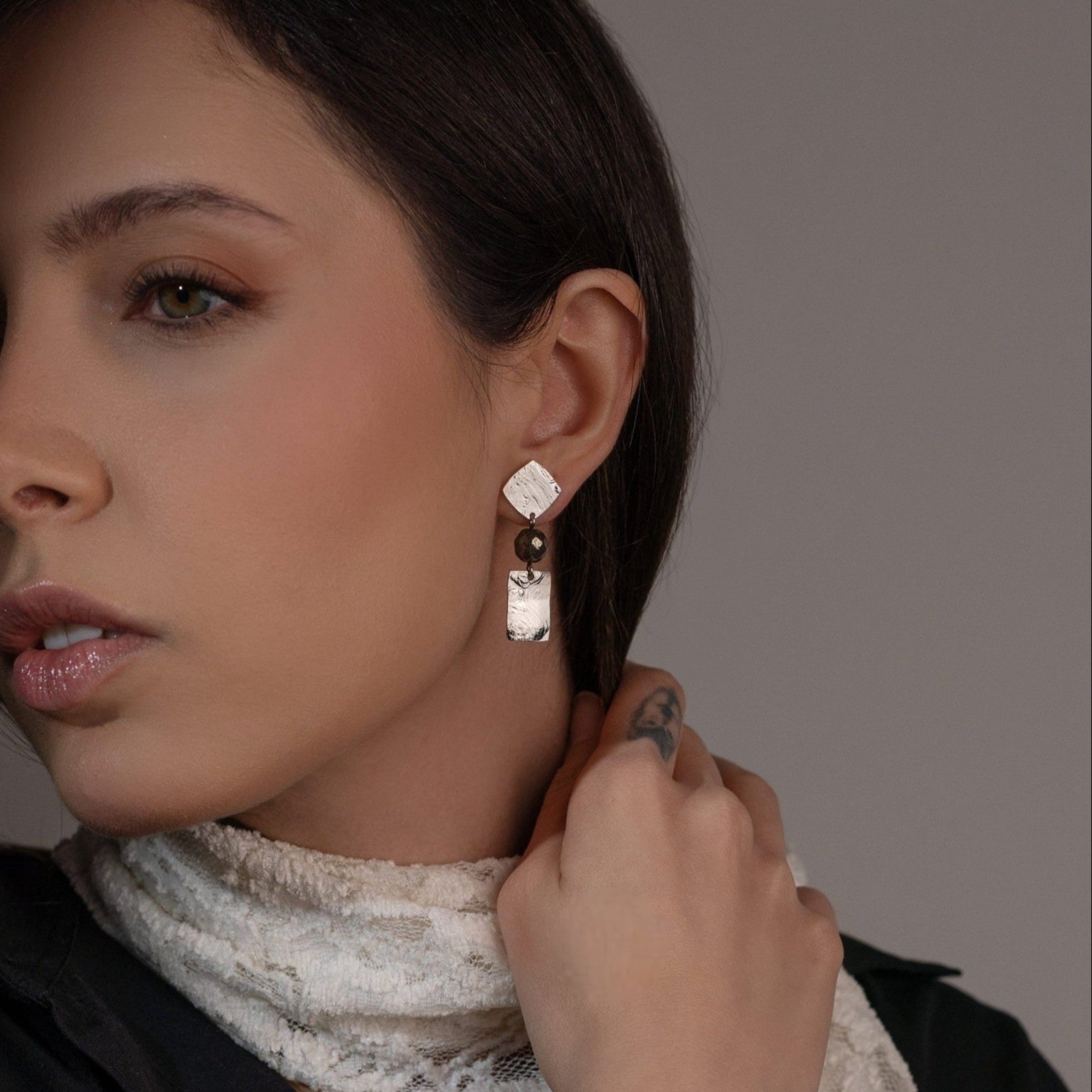 Petites boucles d'oreilles puces en métal martelé argenté et perles de Pyrite, de forme losanges et rectangles, portées à l'oreille par une femme brune