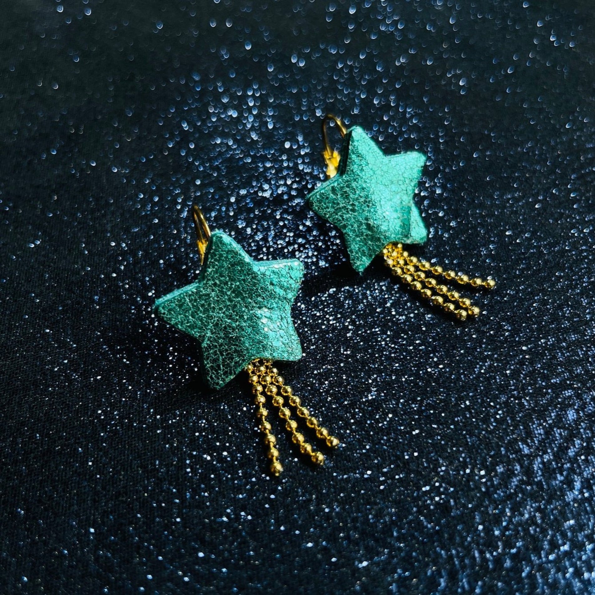 boucles d'oreilles dorées Nune modèle Stella en forme d'étoiles, en cuir vert émeraude brillant intense, sur fond noir paillettes