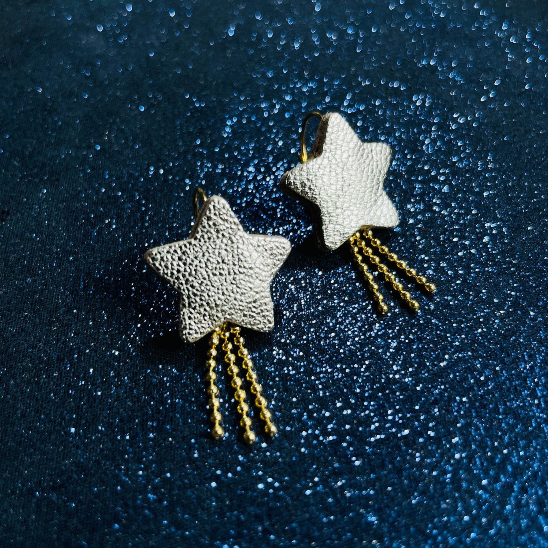 boucles d'oreilles dorées Nune modèle Stella en forme d'étoiles, en cuir argent, sur fond noir paillettes