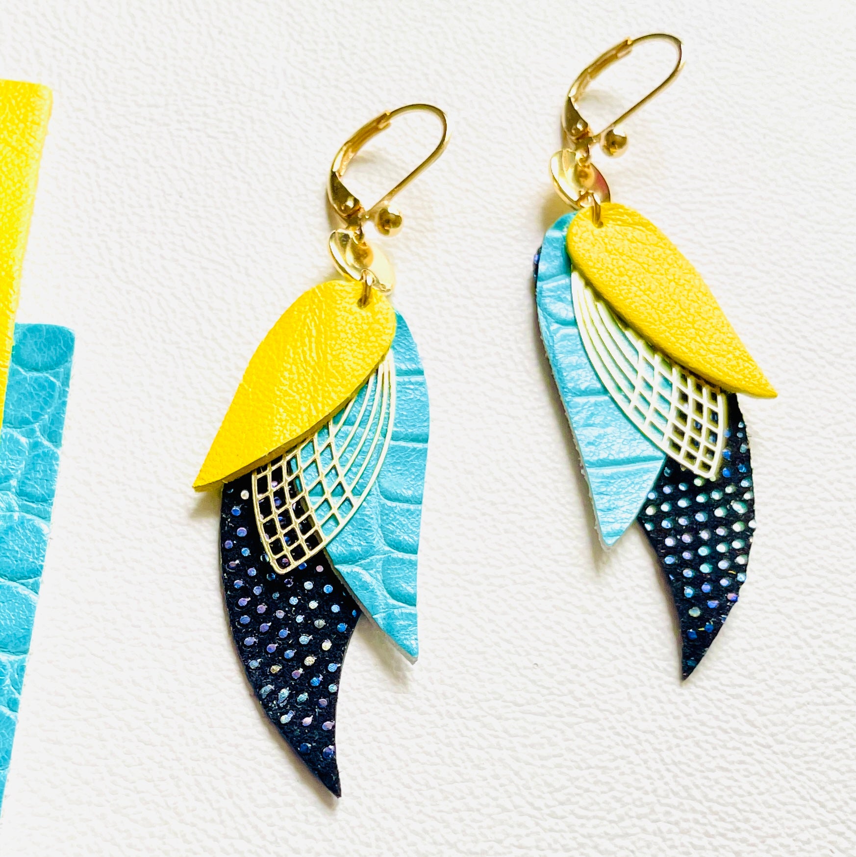 boucles d'oreilles dorées Nune modèle Ara en plumes de cuir jaune citron, turquoise lézard et noir grainé irisé sur fond blanc