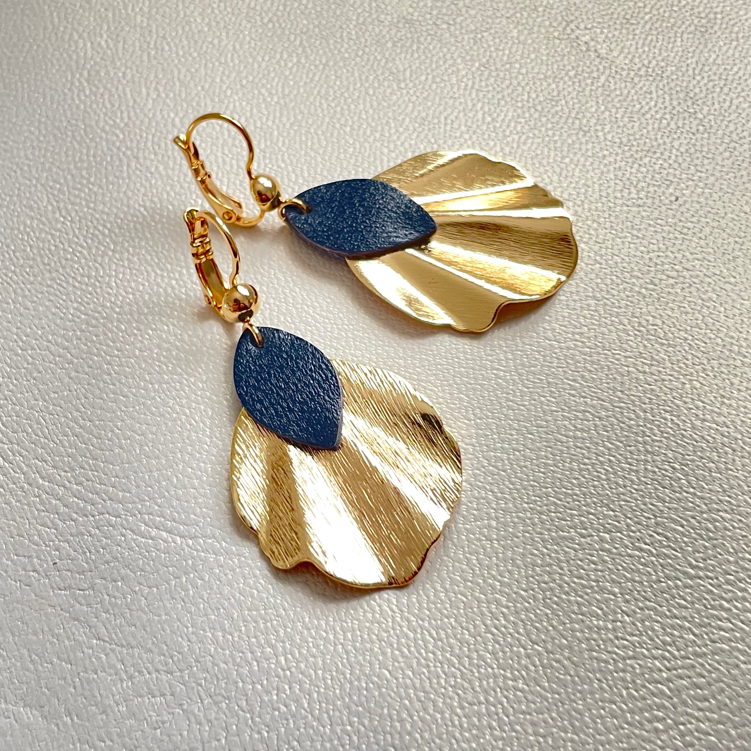 Boucles d'oreilles dorées Nune modÚle Okita en forme de petits coquillages avec pétales de cuir bleu marine satiné, sur fond cuir blanc cassé nacré
