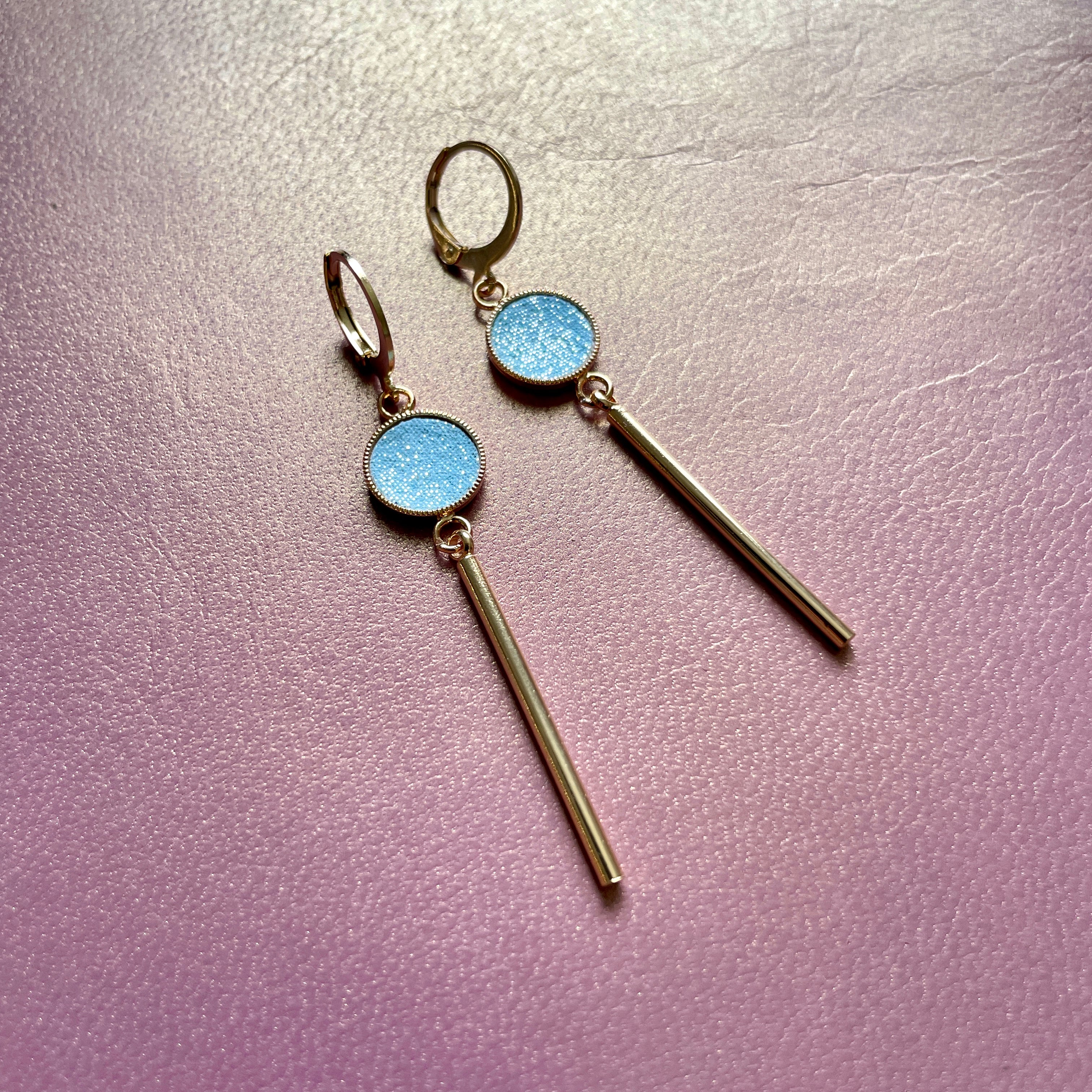 Nune longues et fines boucles d'oreilles dorées Nida avec serti en cuir topaze pailleté, sur fond cuir mauve irisé