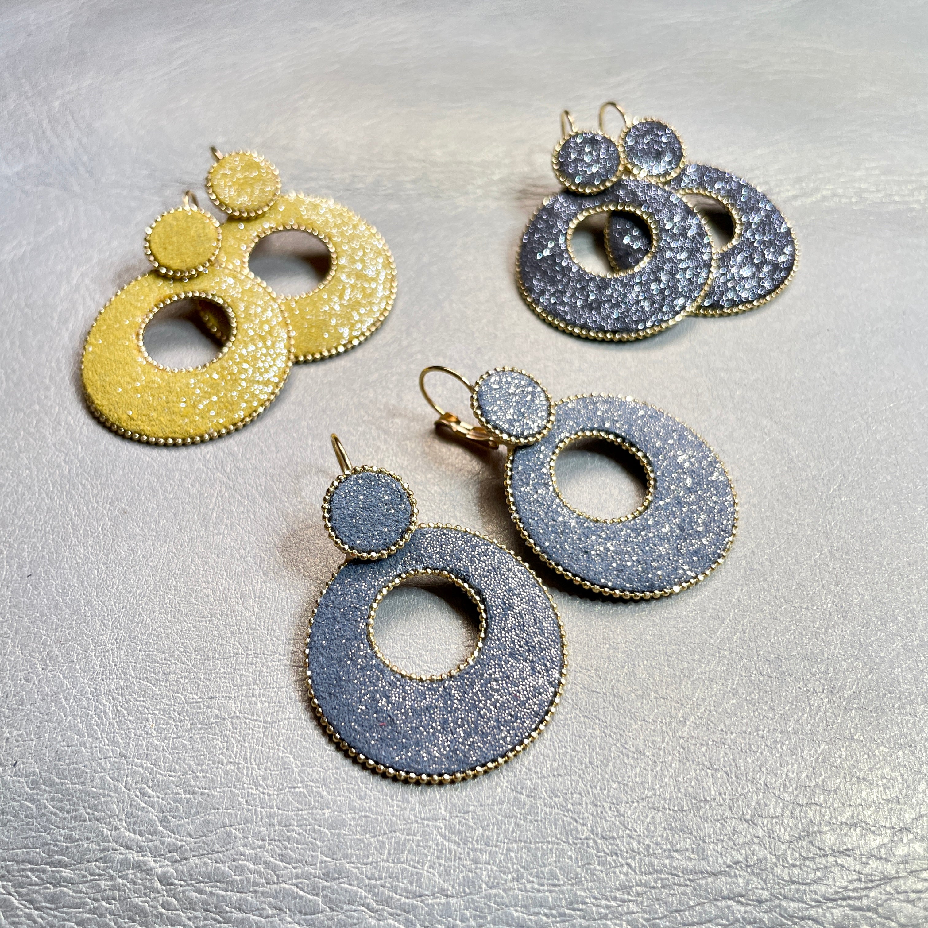 boucles d'oreilles créoles Nune modèle Céleste en forme de cercles en cuir gris pailleté or, safran pailleté argent et anthracite pailleté argent cerclées de chaîne dorée, sur fond beige doré