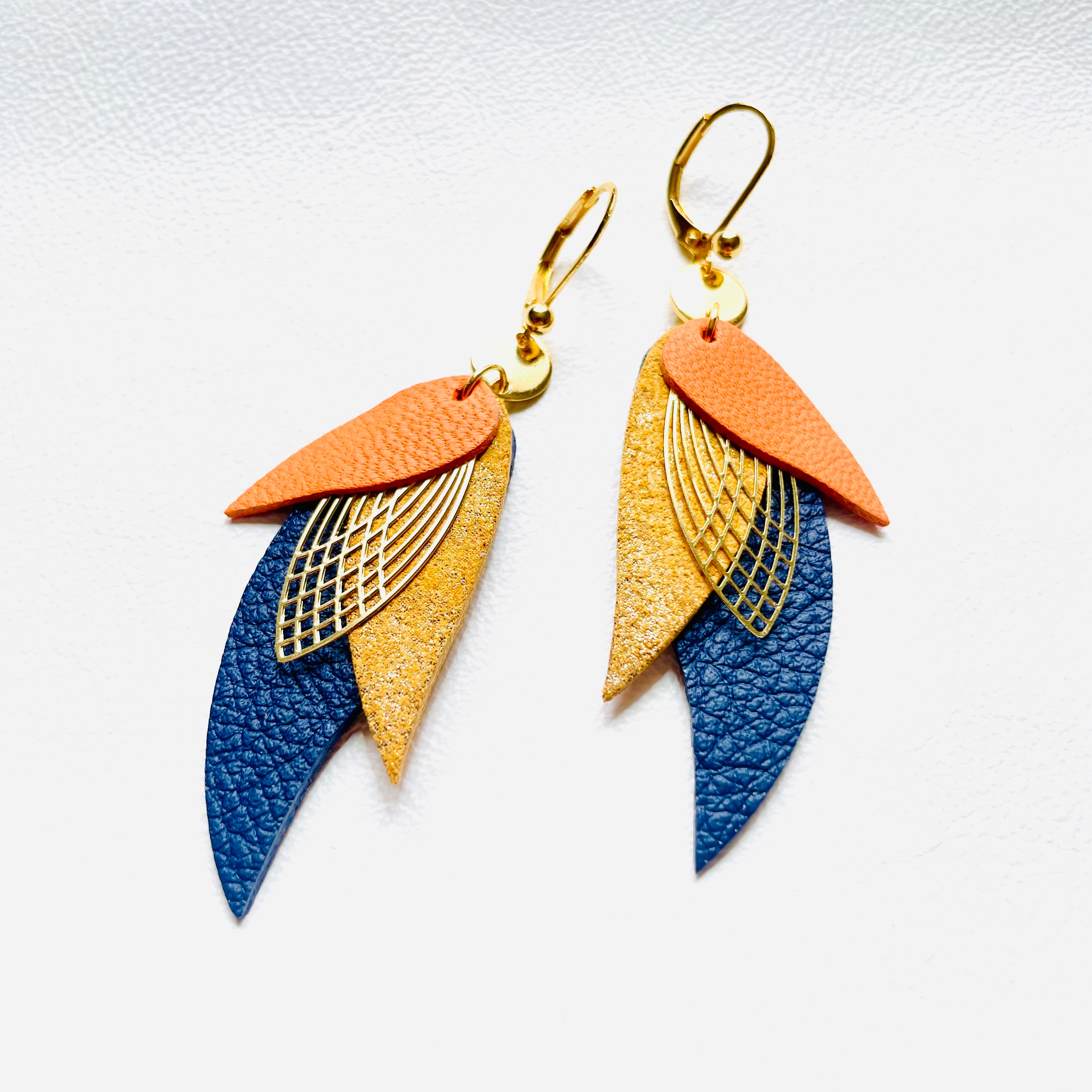boucles d'oreilles dorées Nune modèle Ara en plumes de cuirs abricot, sable doré et bleu marine grainé sur cuir blanc
