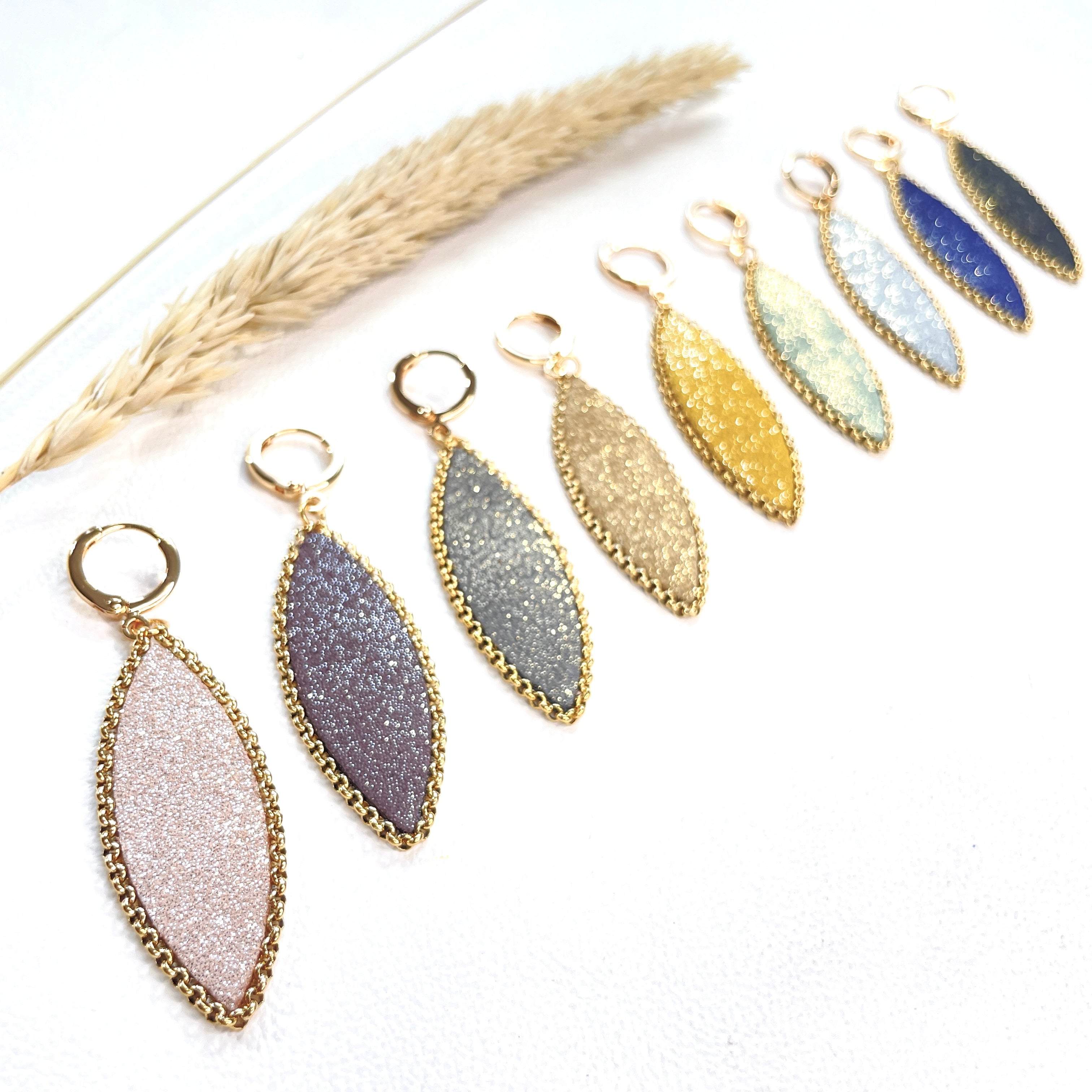 Boucles d'oreilles pendantes Nune modèle Auris en forme d'épi dans 9 coloris de cuirs pailletés cerclés de chaîne dorée, sur fond blanc avec épi