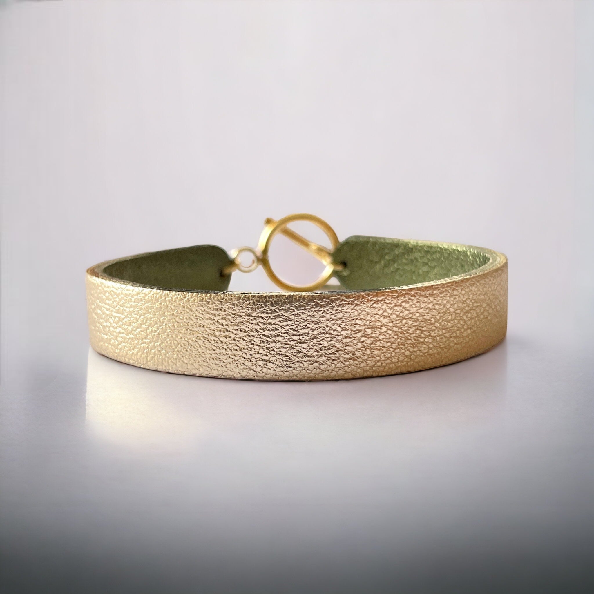 Bracelet en cuir double face bicolore, avec cuir doré antique mat au recto et cuir kaki métallisé au verso avec fermoir boucle dorée en T, sur fond gris