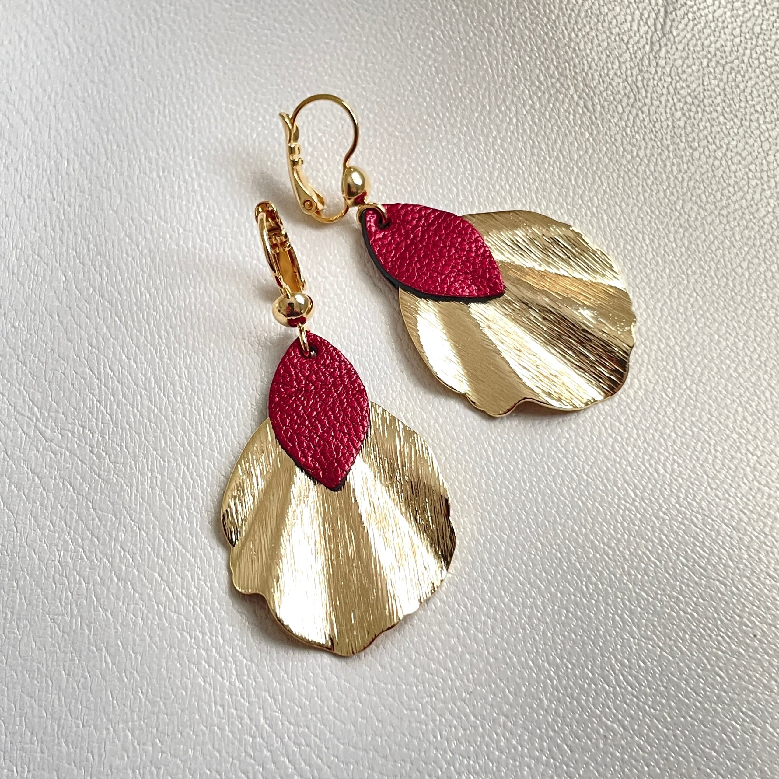 Boucles d'oreilles dorées Nune modÚle Okita en forme de petits coquillages avec pétales de cuir rubis métallisé, sur fond cuir blanc cassé nacré