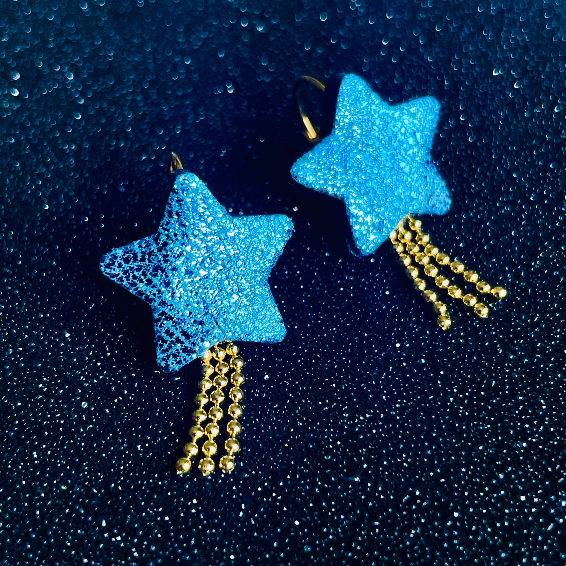 boucles d'oreilles dorées Nune modèle Stella en forme d'étoiles, en cuir bleu roi métallisé, sur fond noir paillettes