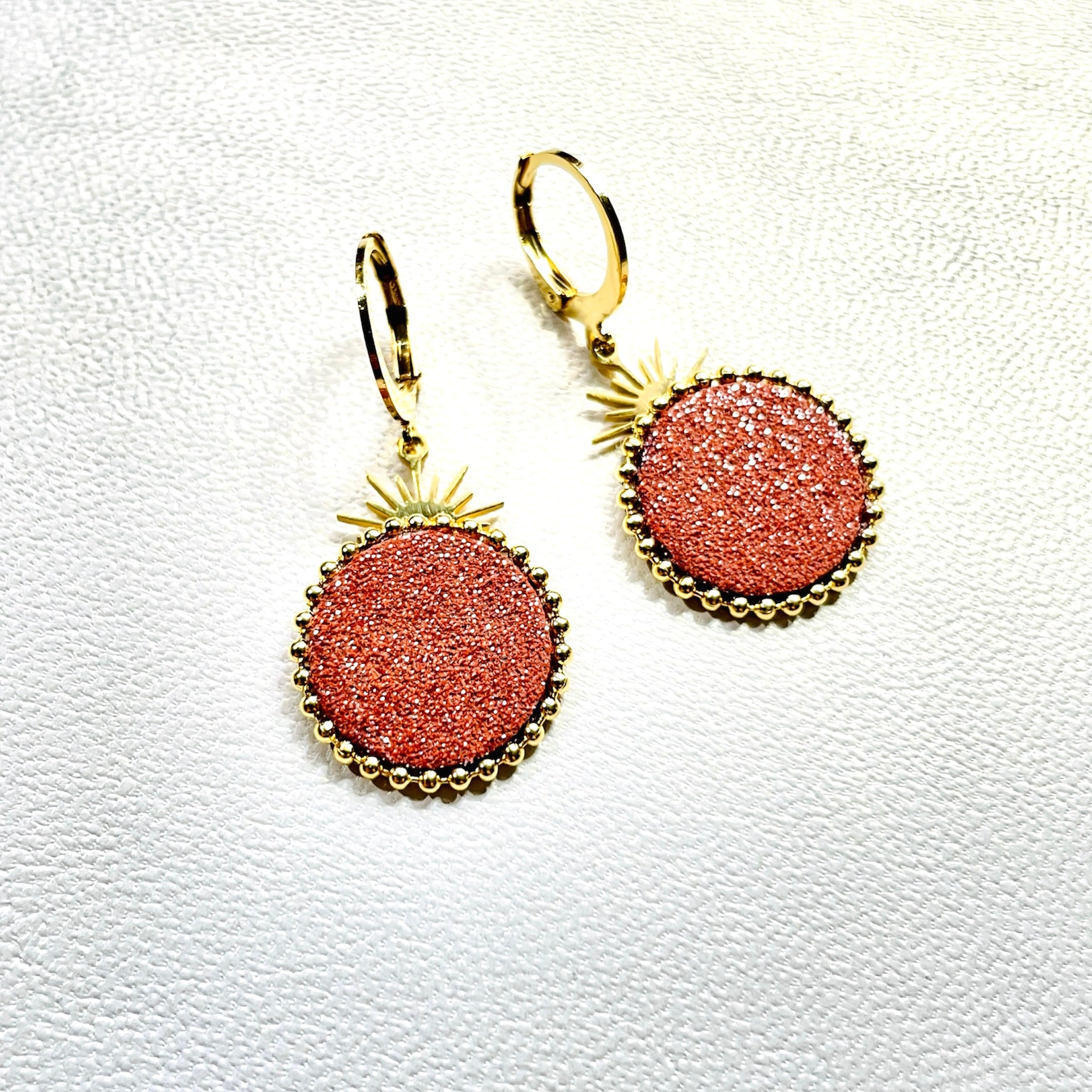 Petites boucles d'oreilles Nune modèle Enia en forme de soleils en cuir pailleté brique pailleté argent, cerclés de chaîne dorée, sur fond blanc