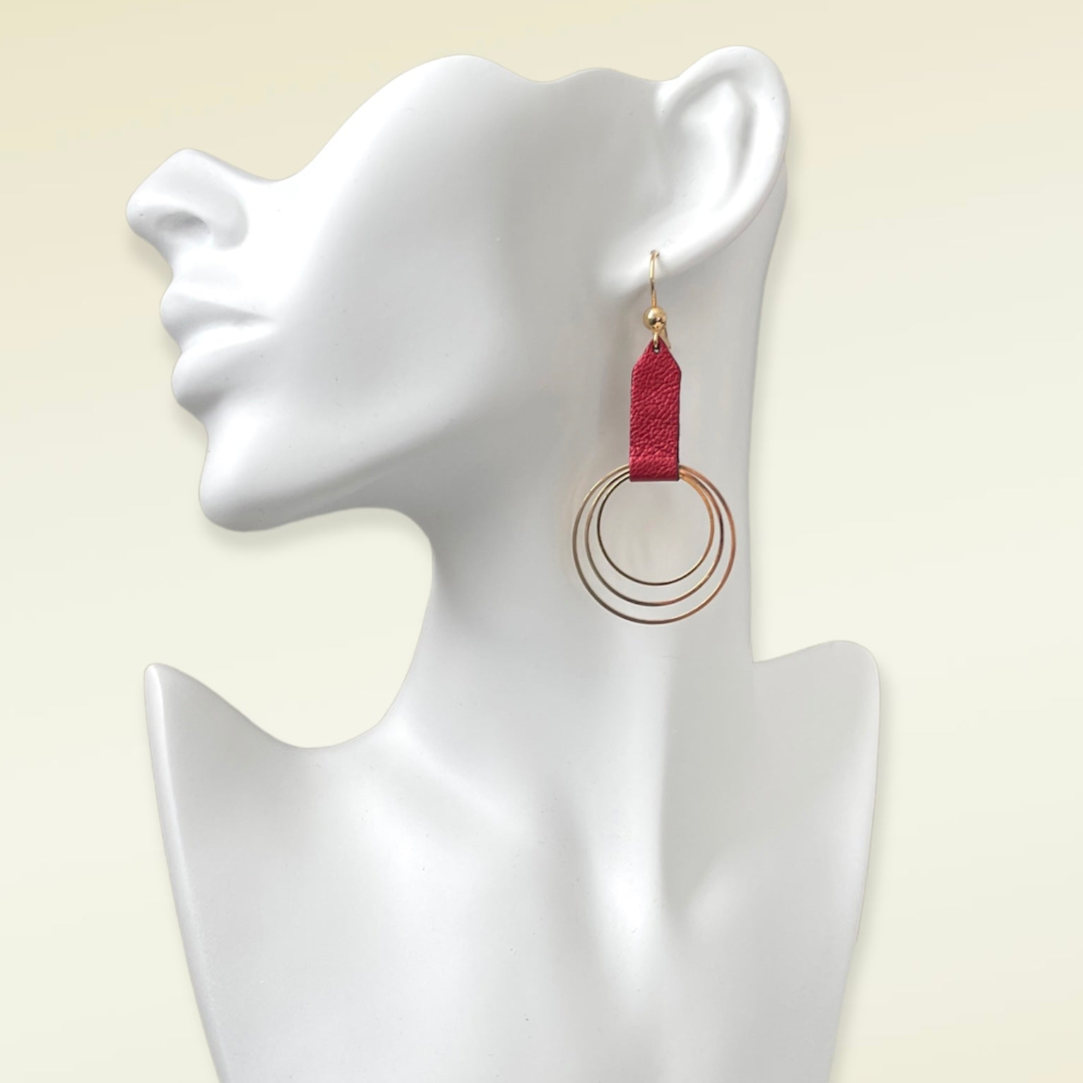 Grande boucle d'oreille dorée avec pendentif en anneaux concentriques attaché par un ruban de cuir recyclé rubis métallisé, portée à l'oreille. Modèle Dizzo