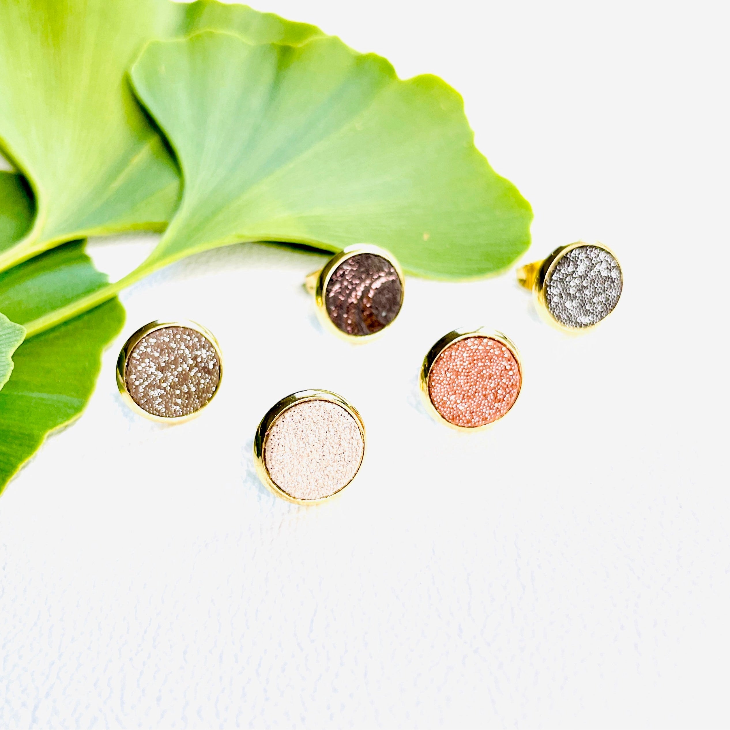 Puces d'oreilles dorées rondes Nune modèle Ilis dans 5 coloris de cuirs pailletés rosés, sur fond blanc avec feuilles de Gingko