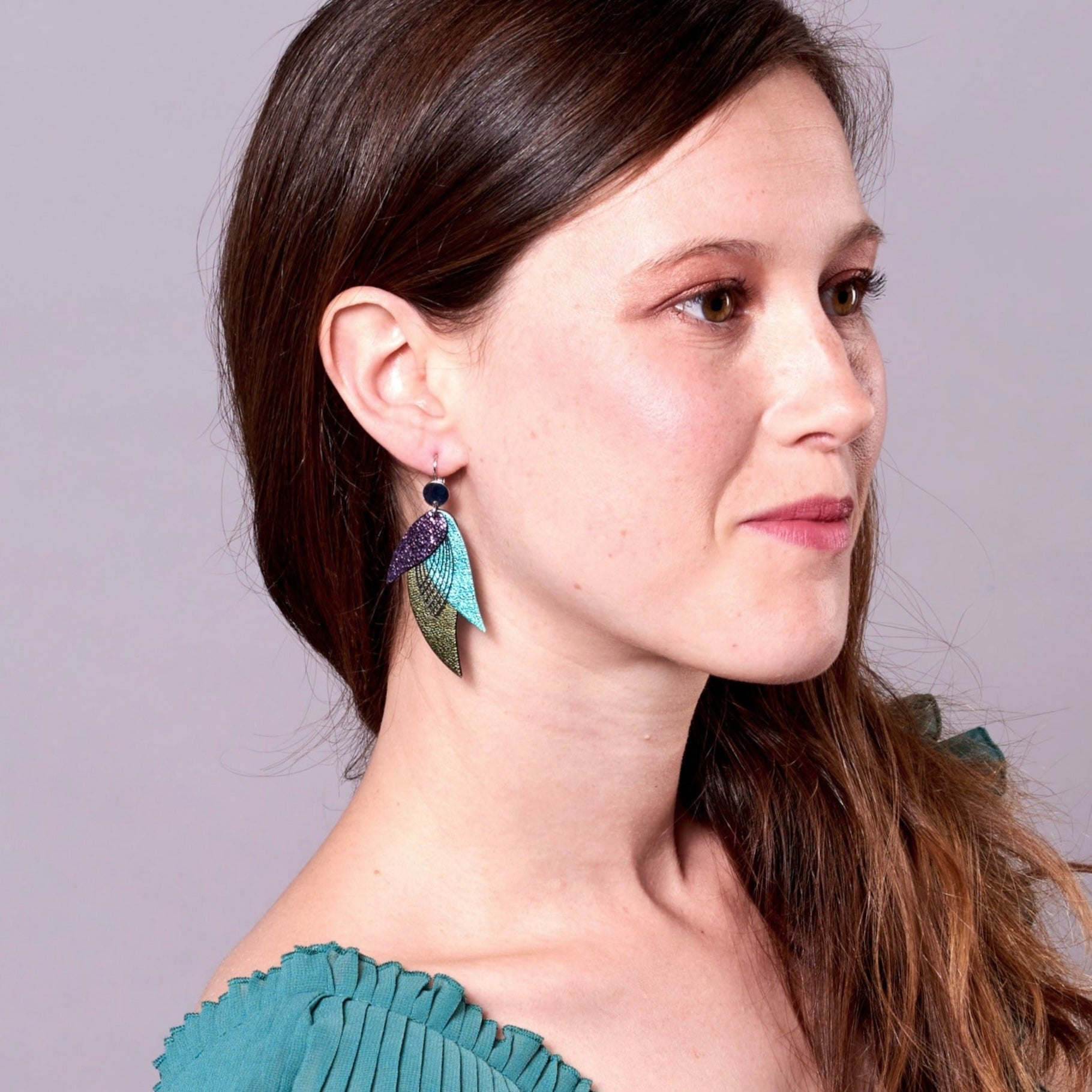 Jeune femme vue de profil portant les Boucles d'oreilles argentées Nune en forme de plumes, modèle Ara en cuirs violet brillant façon serpent, turquoise métallisé et kaki métallisé sur fond mauve