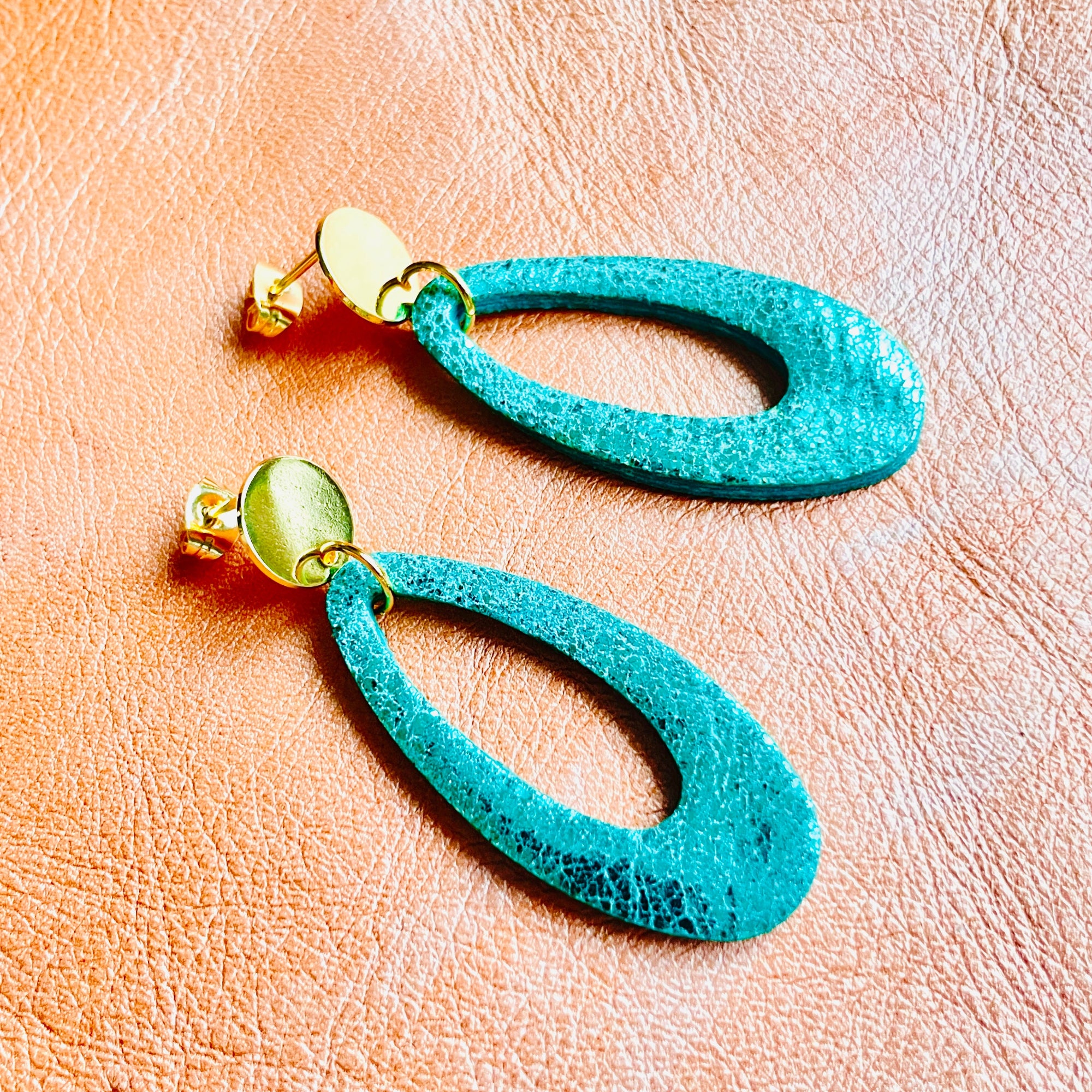 Puces d'oreilles dorées Drys de la marque Nune, avec leurs anneaux ovales en cuir vert émeraude brillant, sur fond cuir corail nacré clair, vues de côté