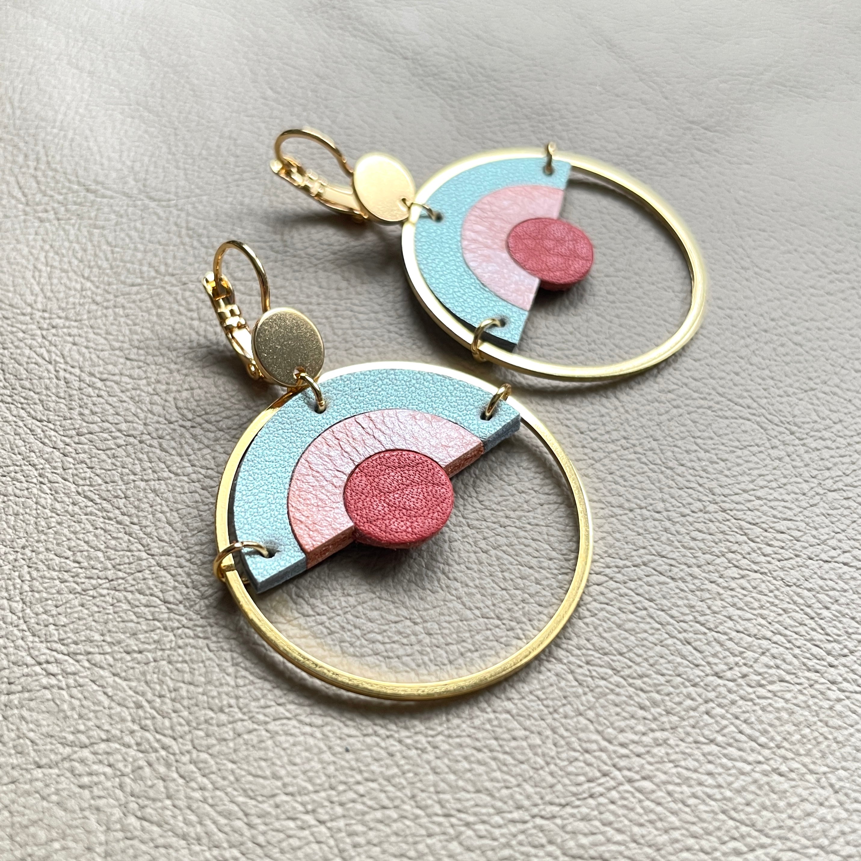 Boucles d'oreilles dorées et rondes, modèle Sunset de la marque Nune, avec un dégradé arc-en-ciel de cuirs rose incarnat, corail nacré clair et bleu lune nacré, sur fond beige
