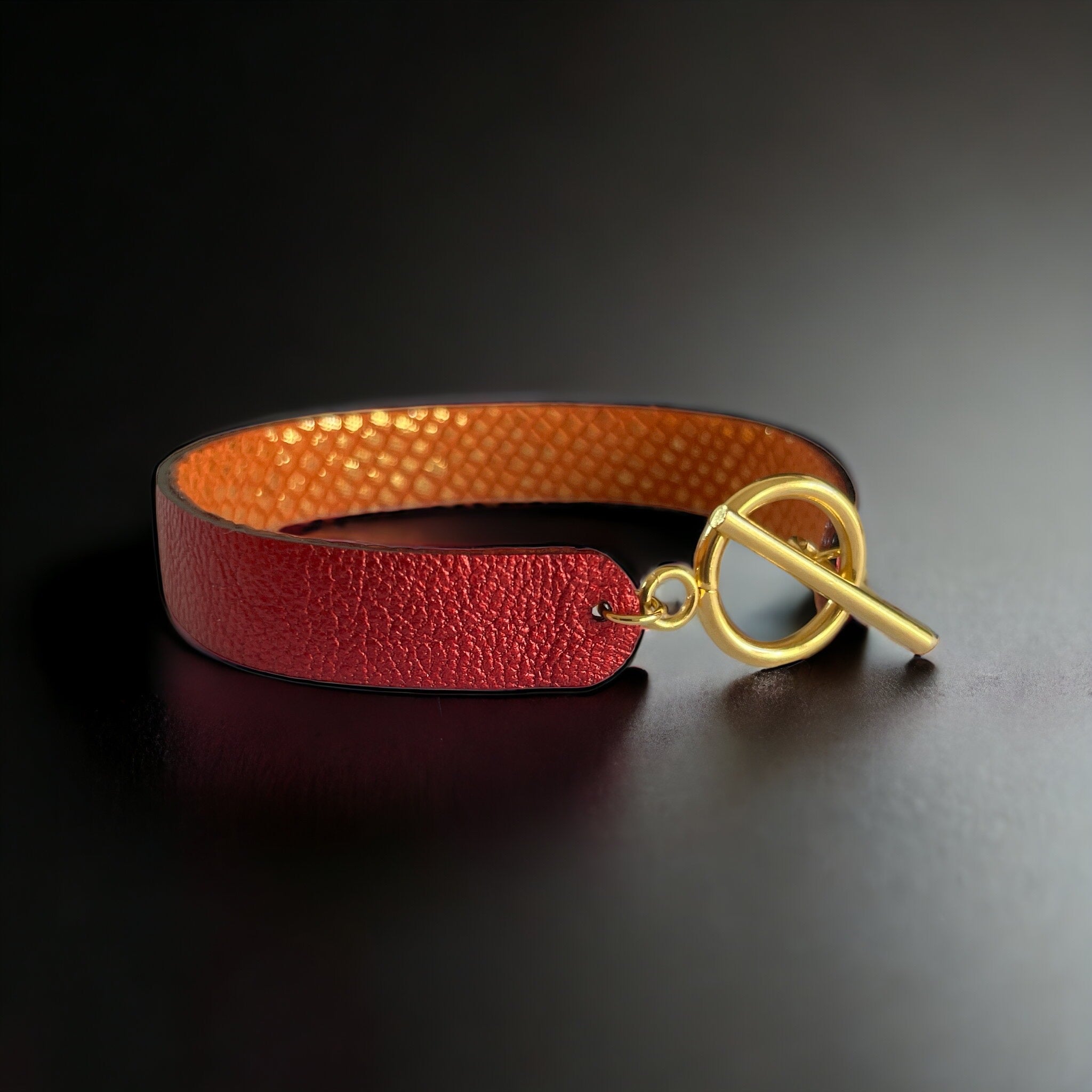Bracelet en cuir double face bicolore, avec cuir rubis métallisé au recto et cuir orangé or façon croco au verso avec fermoir boucle dorée en T, sur fond noir