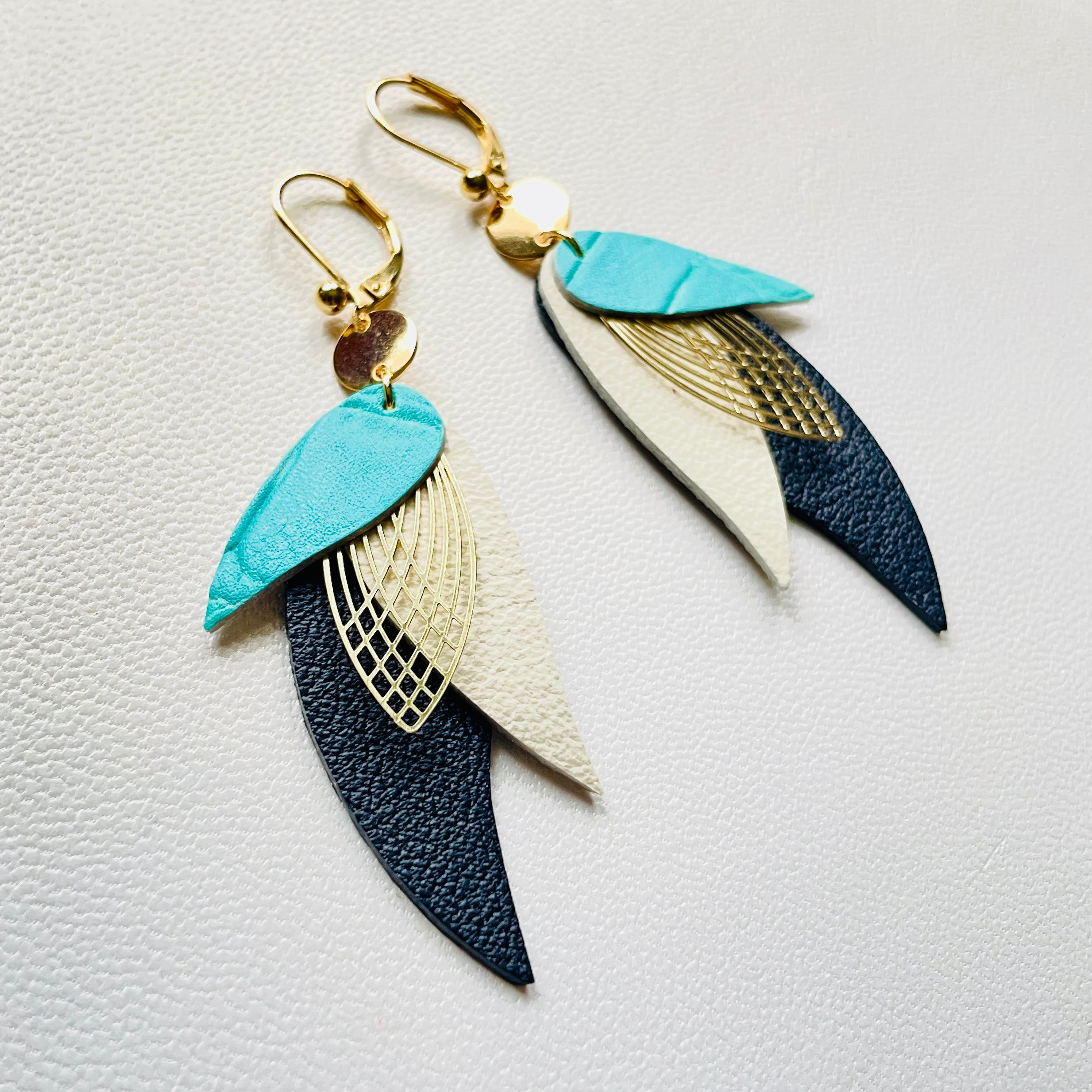 boucles d'oreilles dorées Nune modèle Ara en plumes de cuirs turquoise lézard, blanc cassé mat et noir grainé sur cuir blanc