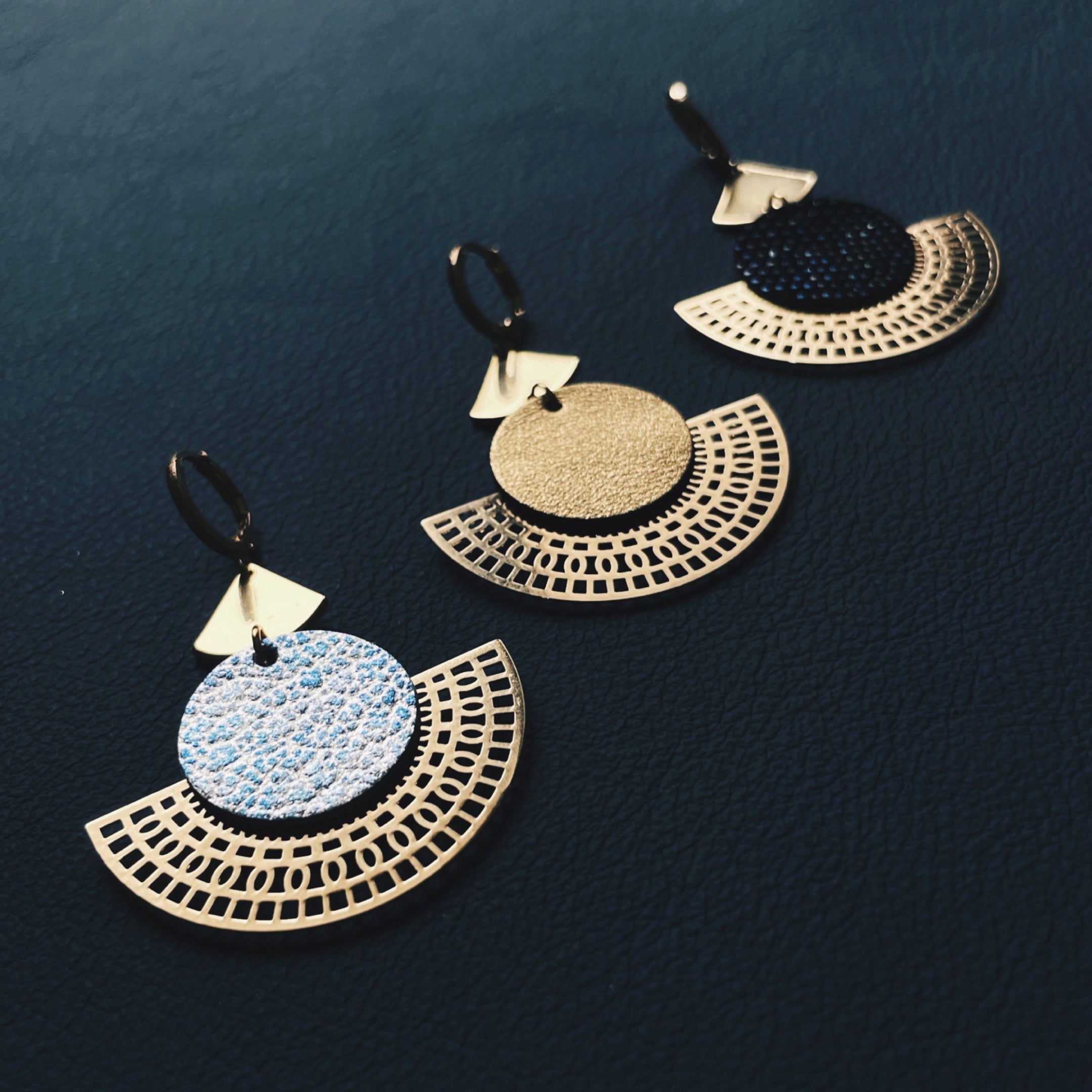 Boucles d'oreilles dorées Nune modèle Ephésia en cuir bleu ciel métal patiné, doré satiné et noir grainé irisé sur fond noir
