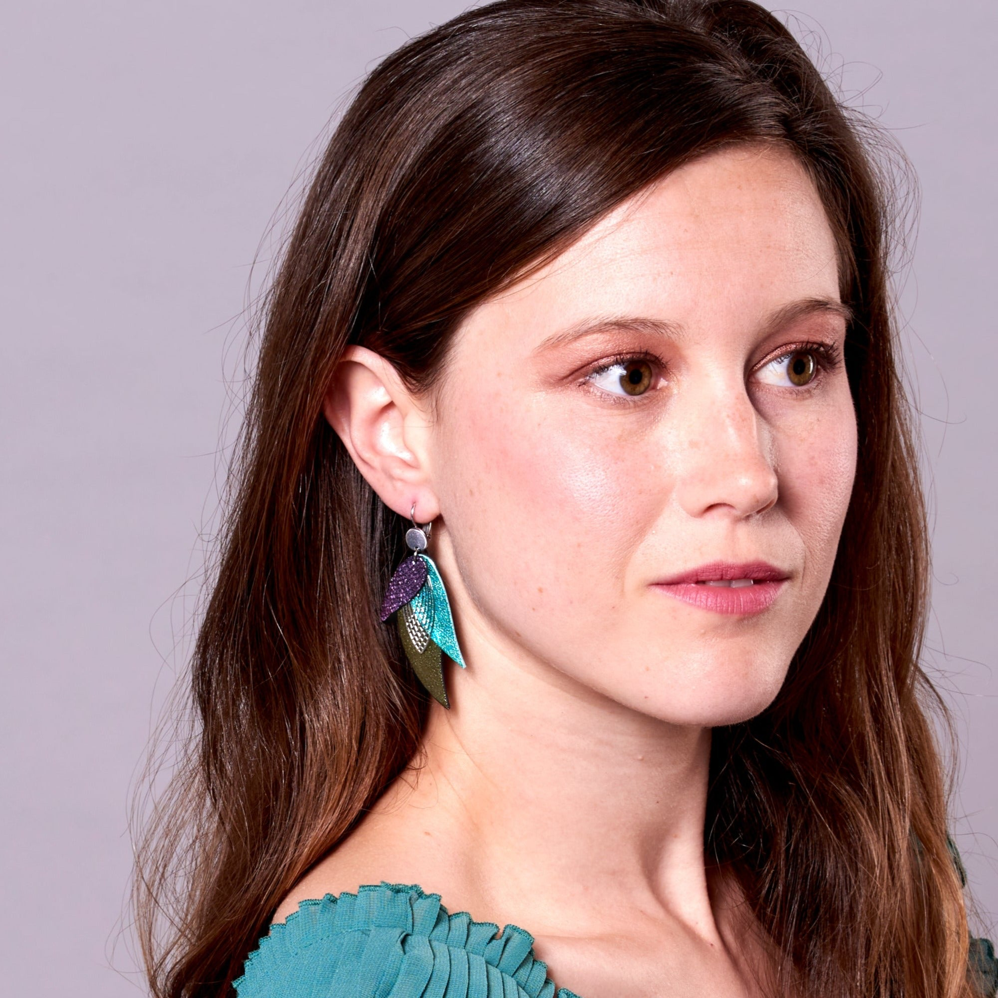 Jeune femme portant les Boucles d'oreilles argentées Nune en forme de plumes, modèle Ara en cuirs violet brillant façon serpent, turquoise métallisé et kaki métallisé sur fond mauve