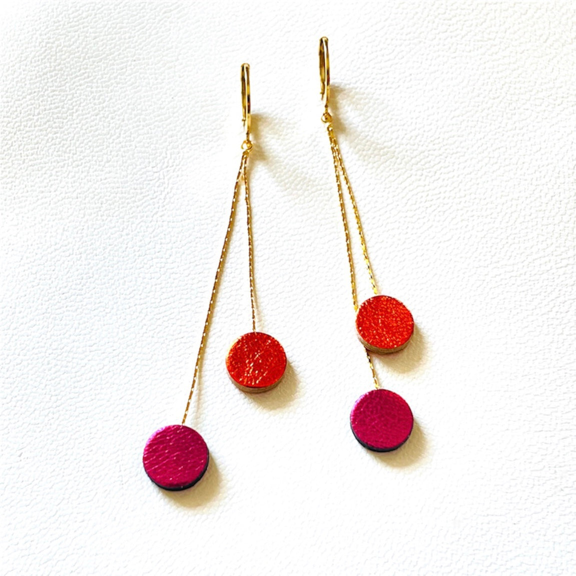 Longues et fines boucles d'oreilles Nune dorées terminées petits disques de cuir recyclé orange sanguine métallisé et fushia métallisé, sur fond blanc. Modèle Merry