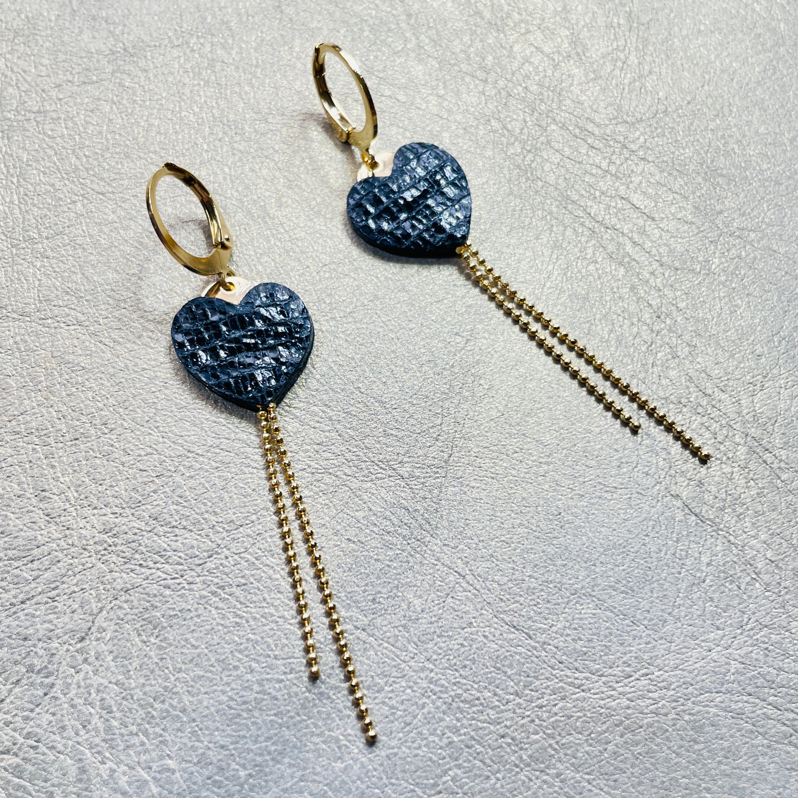 NUNE fines boucles d'oreilles dorées, petits cœurs de cuir recyclé noir brillant façon serpent et chaînettes, sur fond gris