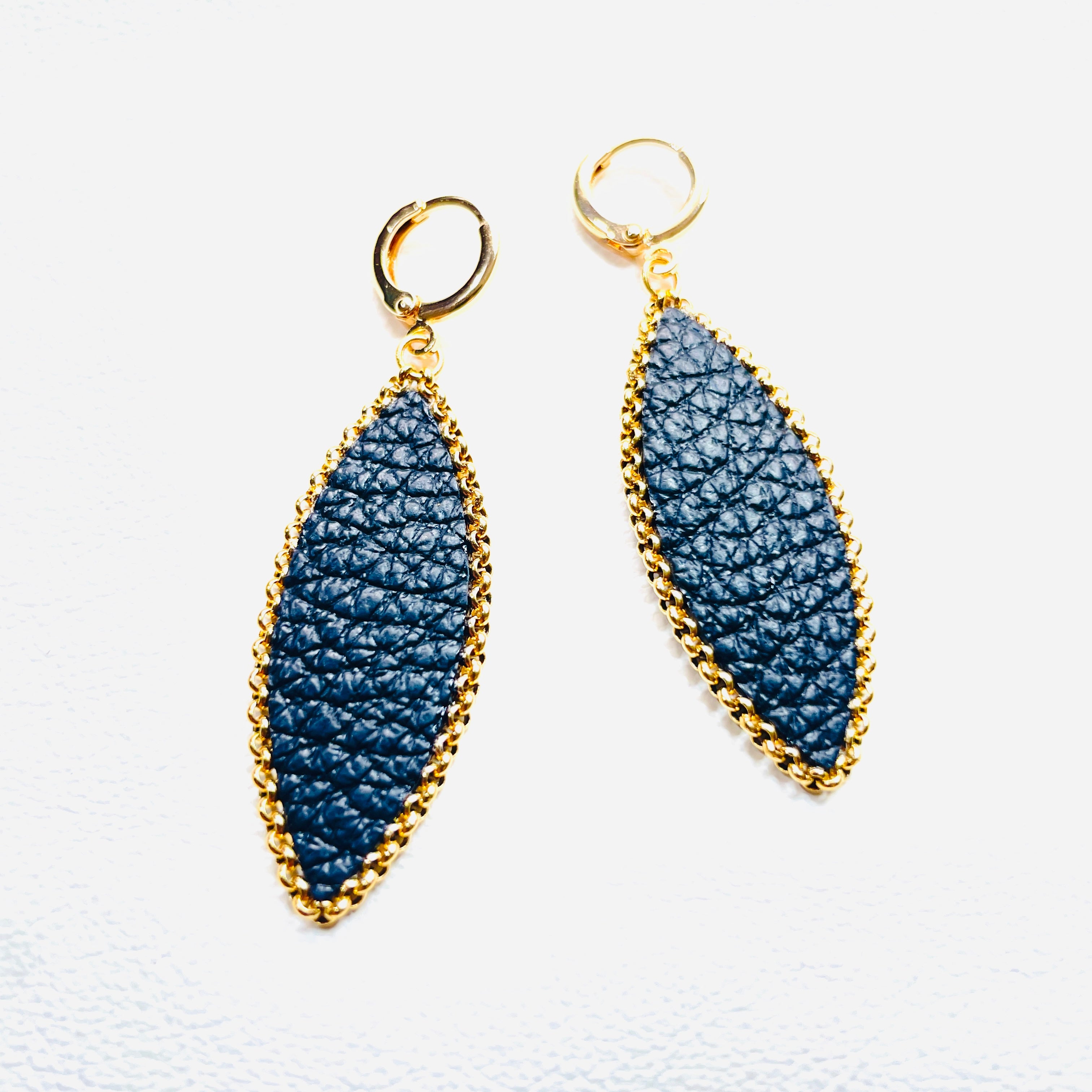 Paire de boucles d'oreilles pendantes Nune modèle Auris en forme d'épi en cuir noir grainé cerclé de chaîne dorée, sur fond cuir blanc