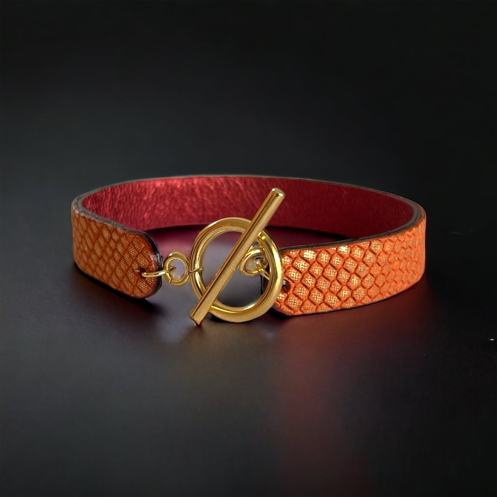 Bracelet en cuir double face bicolore, avec cuir rubis métallisé au verso et cuir orangé or façon croco au recto avec fermoir boucle dorée en T, sur fond noir