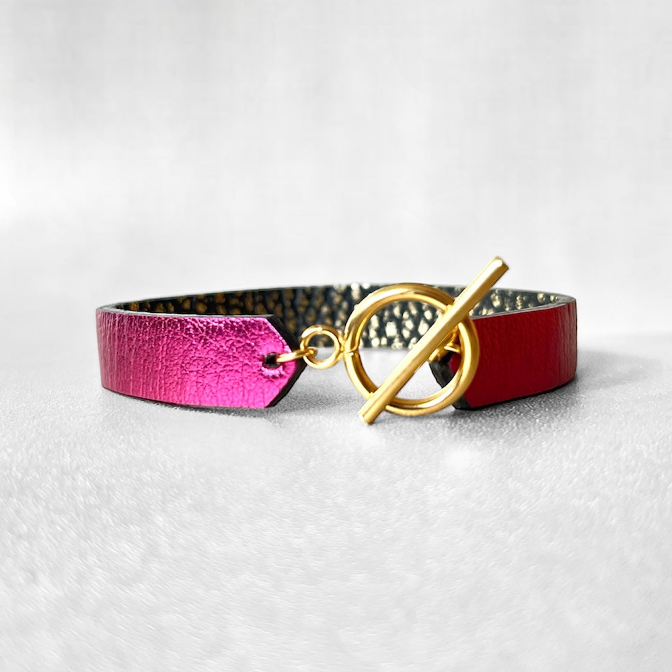 Bracelet en cuir double face bicolore, avec cuir fushia métallisé au recto et cuir noir grainé or au verso avec fermoir boucle dorée en T, sur fond gris