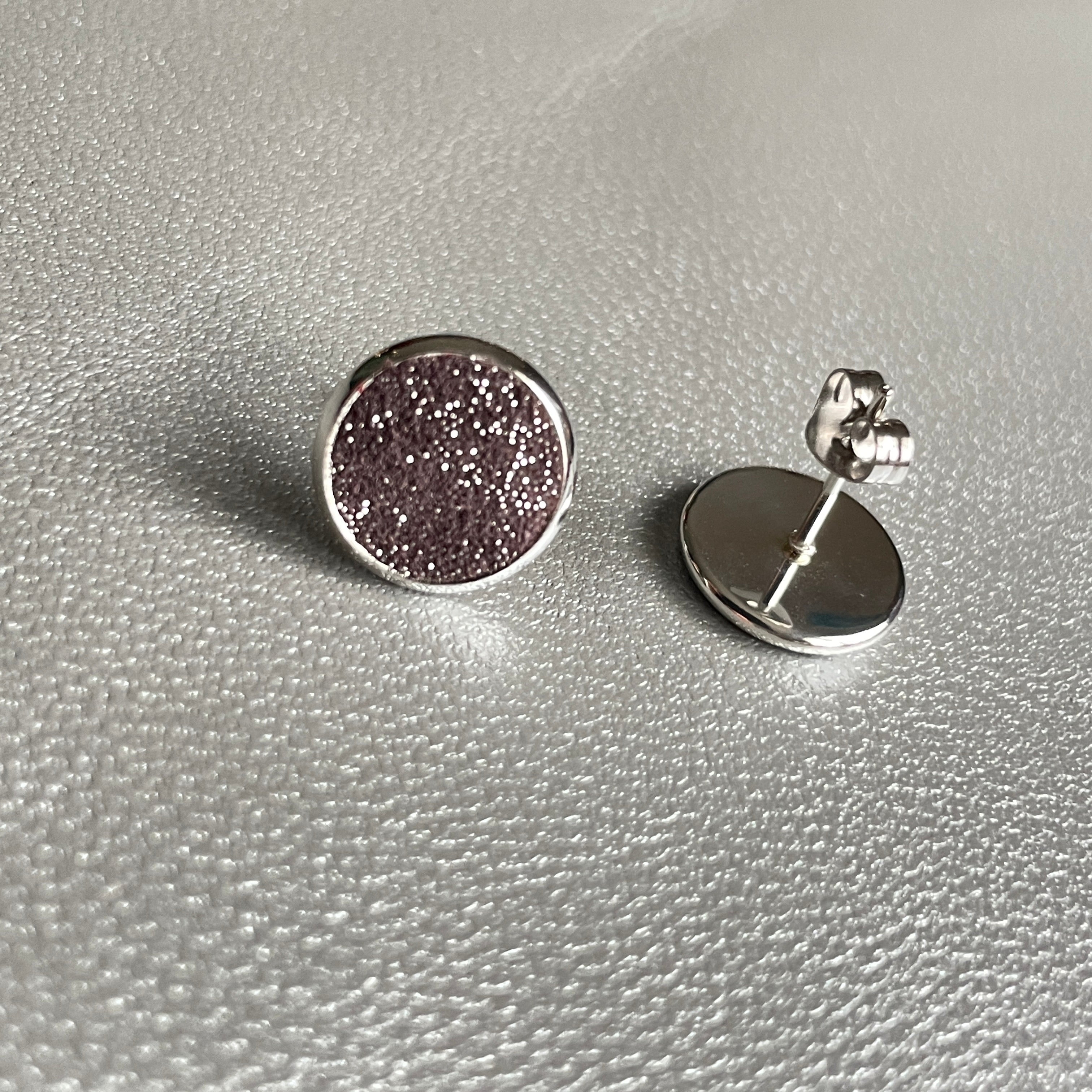 Puces d'oreilles argentées rondes Nune modèle Ilis argenté pailleté avec pastilles de cuir parme pailleté argent, sur fond de cuir gris métallisé