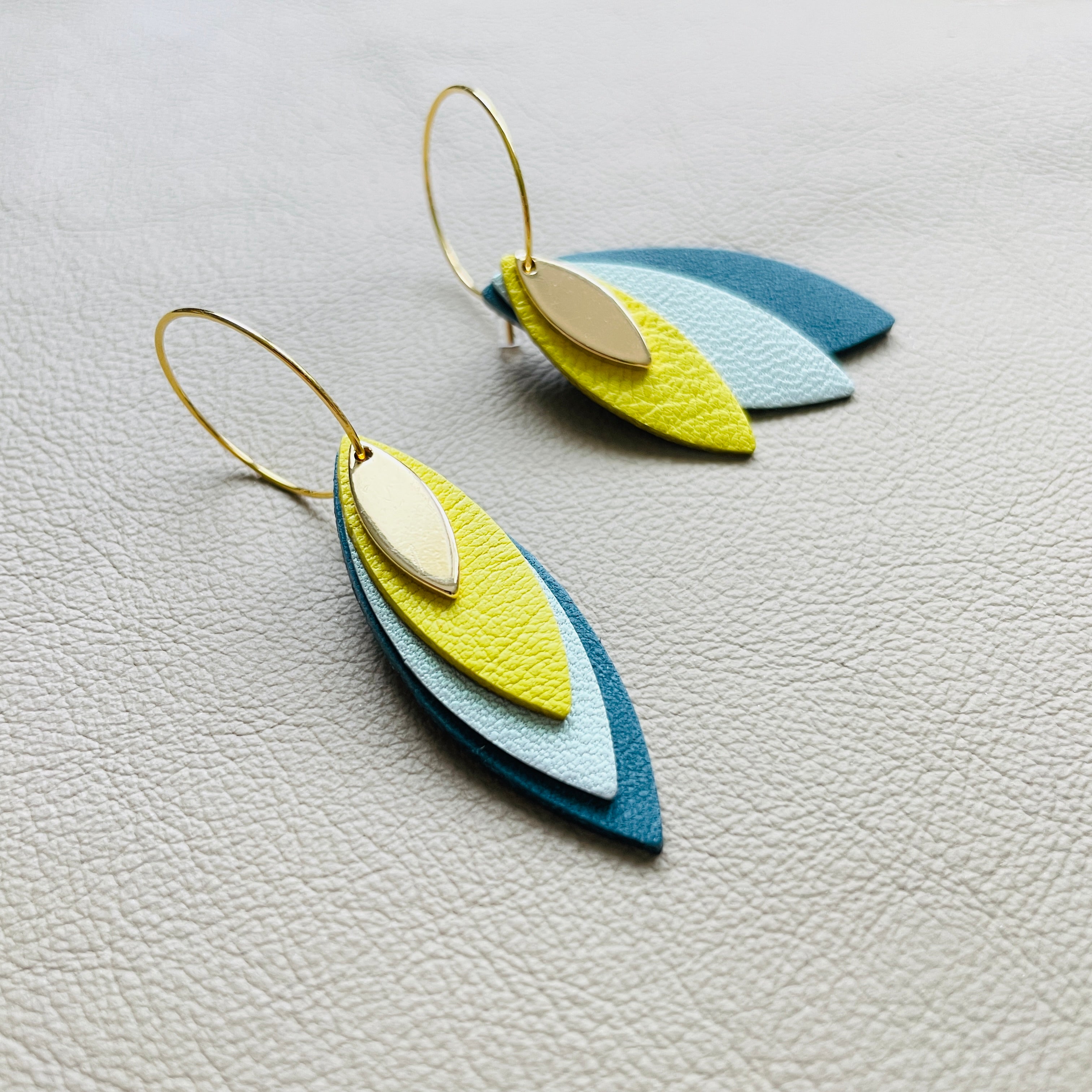 Boucles d'oreilles créoles dorées Nune modèle Piume en cuirs jaune citron, bleu lune nacré et vert paon sur fond gris